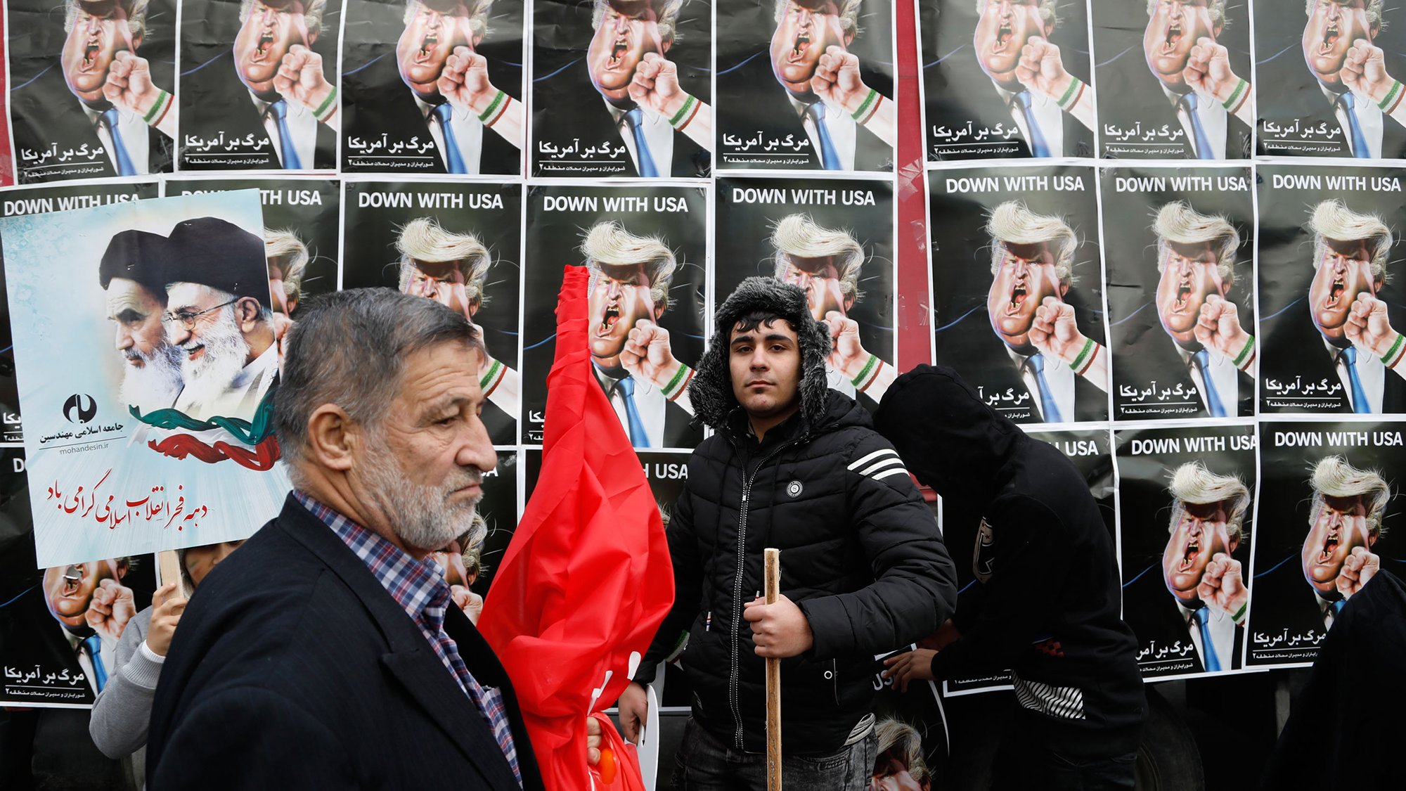L'image montre une manifestation avec des affiches en arrière-plan affichant des personnages aux visages provocateurs, probablement des figures politiques, portant des coiffures spécifiques. Des gens tiennent des pancartes et des drapeaux, tandis qu'un homme sur le devant semble exprimer un message clair de désaccord ou de protestation. L'ensemble de la scène dégage une ambiance de contestation, soulignant des tensions politiques.
