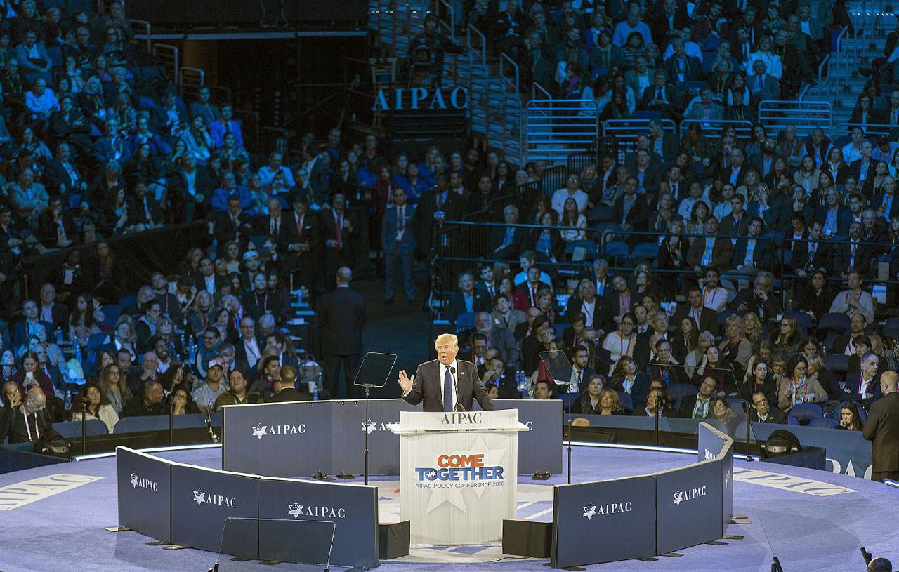 L'image montre un grand rassemblement dans une salle pleine de spectateurs. Au centre, une scène où un orateur, probablement un homme politique, s'exprime devant le public. Le logo "AIPAC" est visible sur des panneaux autour de la scène, indiquant que cet événement est lié à l'organisation pro-israélienne. Les participants semblent attentifs, et l'ambiance générale évoque un moment solennel et engagé. Les couleurs de l'éclairage sont principalement bleu et violet.