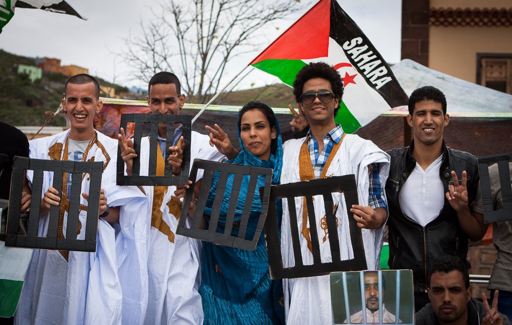 L'image montre un groupe de personnes posant ensemble lors d'une manifestation. Ils portent des vêtements traditionnels, notamment des robes blanches. Certains affichent des signes pacifiques avec leurs mains, tandis que d'autres tiennent des panneaux représentant des barreaux de prison. En arrière-plan, on peut voir un drapeau aux couleurs du Sahara. L'atmosphère semble engagée et solidaire.