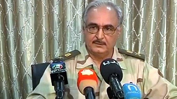 L'image montre un homme portant un uniforme militaire avec des rangs. Il est assis devant plusieurs microphones, suggérant qu'il est en train de donner une déclaration ou une interview. L'arrière-plan est composé de rideaux clairs, et l'homme a une expression sérieuse. On sent qu'il s'agit d'une situation officielle.