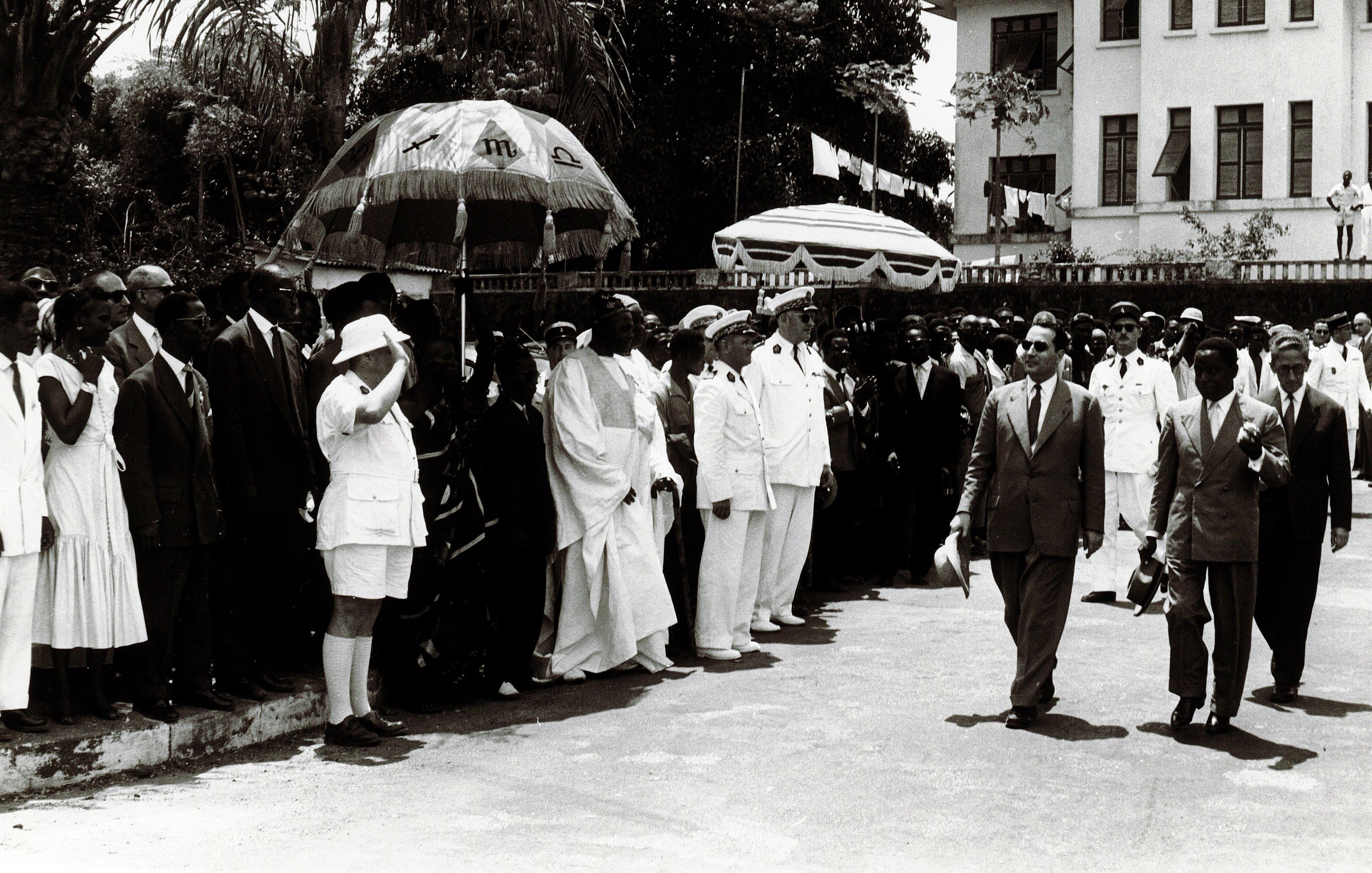 L'image montre une scène publique, probablement une cérémonie officielle. On y voit un groupe de personnes en costume, regroupées sur un côté, avec des hommes portant des chapeaux et des vêtements formels. À l'avant, on distingue des hommes en uniformes militaires et d'autres en tenue élégante. Au centre, une figure principale semble marcher vers la caméra, entourée de gardes et d'accompagnateurs. Des parasols colorés sont également visibles, ajoutant une touche d'ombre à la scène. En arrière-plan, on peut apercevoir un bâtiment officiel avec des drapeaux, indiquant un événement important ou une célébration. L'atmosphère semble solennelle et organisée.