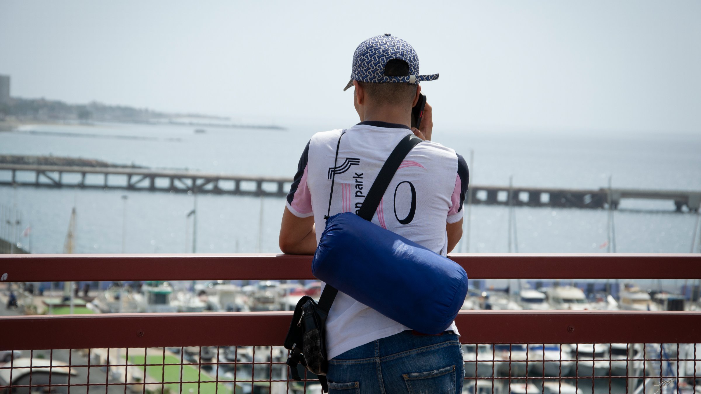 Un homme regarde la mer depuis un pont, avec un port et des bateaux en arrière-plan.