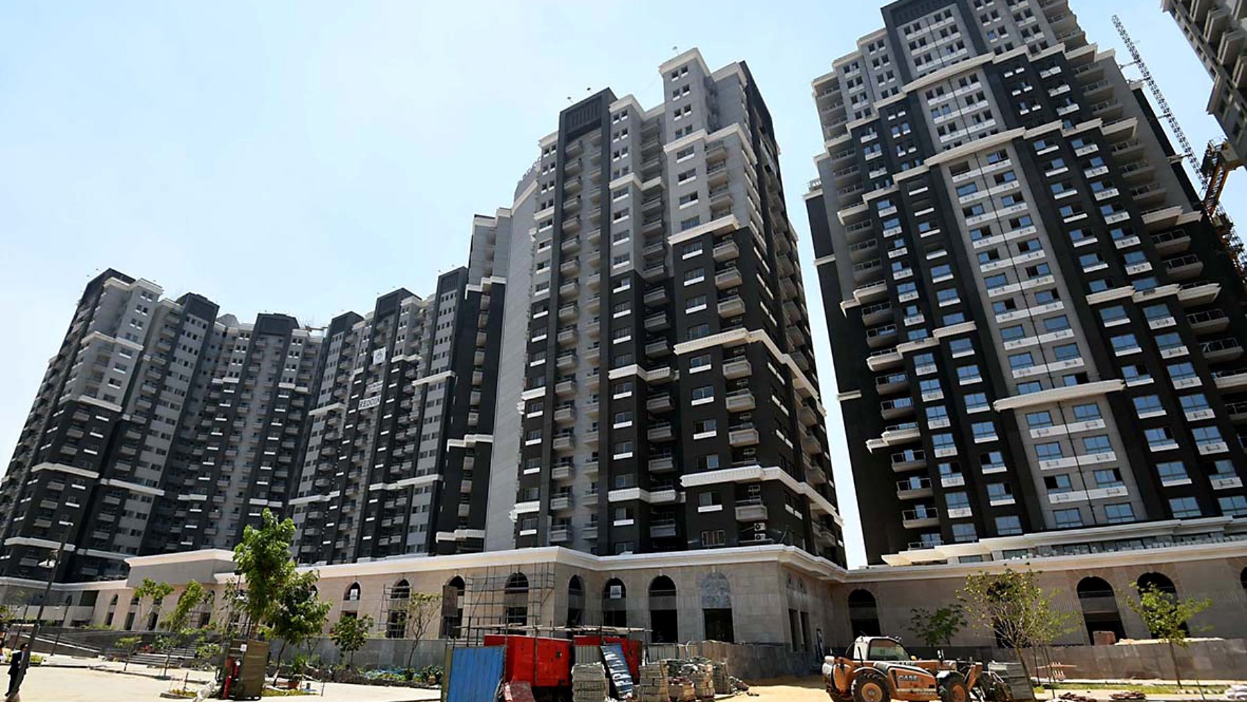 L'image montre un ensemble de grands immeubles résidentiels modernes. Les bâtiments sont principalement de couleur noire et blanche, avec de grandes fenêtres qui laissent entrer la lumière. On peut voir un chantier en cours au premier plan, avec des véhicules de construction et des matériaux dispersés. Il y a aussi quelques arbres et des espaces verts, suggérant un aménagement paysager en développement. Le ciel est dégagé et ensoleillé.