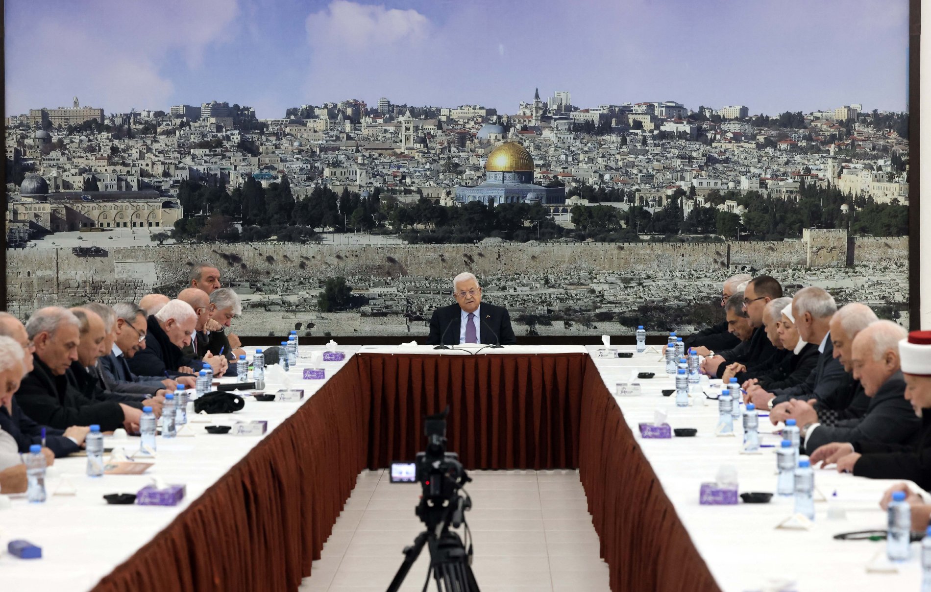 L'image montre une réunion se tenant autour d'une grande table ovale, avec plusieurs personnes assises de chaque côté. Au fond, il y a une grande image d'une ville, probablement Jérusalem, avec le dôme doré d'une mosquée bien visible. Les participants, qui semblent être des hommes âgés, portent des costumes sombres et affichent une expression sérieuse. Au centre de la table, une personne (probablement un chef ou un président) s'exprime pendant que les autres l'écoutent attentivement. Des bouteilles d'eau et des serviettes sont disposées sur la table. L'ambiance semble solennelle et attentive.