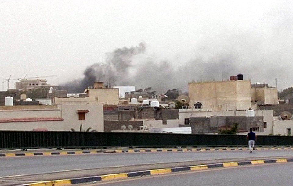 L'image montre une scène urbaine avec des bâtiments de différentes tailles, typiques d'un environnement urbain. Au loin, on peut apercevoir des colonnes de fumée sombre s'élevant dans le ciel, suggérant probablement un incendie ou une explosion. Le ciel est nuageux, ce qui ajoute à l'atmosphère sombre de la scène. Au premier plan, une personne marche le long d'une route, semblant s'éloigner de la source de la fumée. Les couleurs sont plutôt ternes, reflétant une ambiance de tension ou d'urgence.