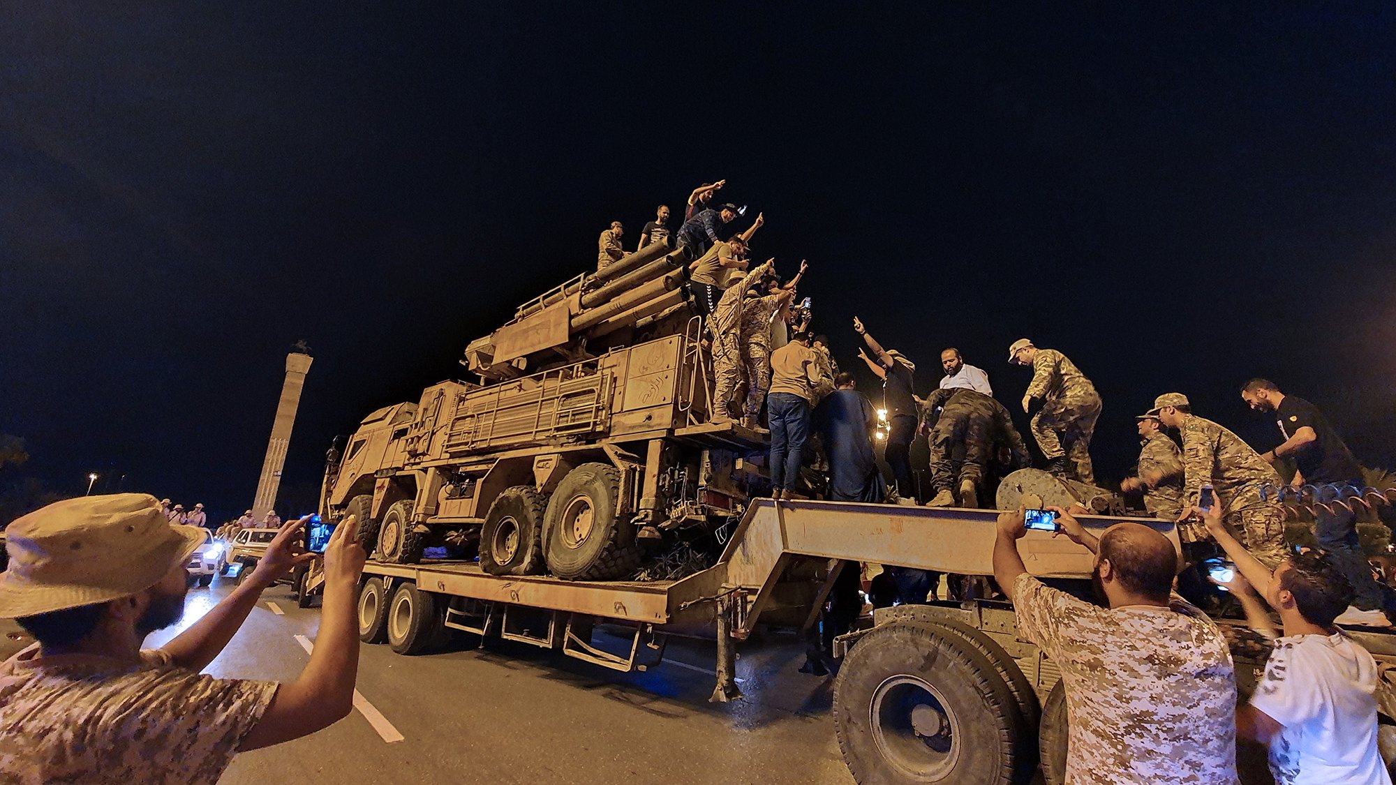 L'image montre un groupe de personnes rassemblées autour d'un véhicule militaire sur une route, probablement pendant la nuit. Plusieurs individus sont montés sur le véhicule et semblent exprimer des émotions fortes, peut-être en célébrant ou en manifestant un soutien. D'autres personnes prennent des photos avec leurs téléphones. L'ambiance générale semble dynamique et impliquée, avec une forte présence militaire sur le site.