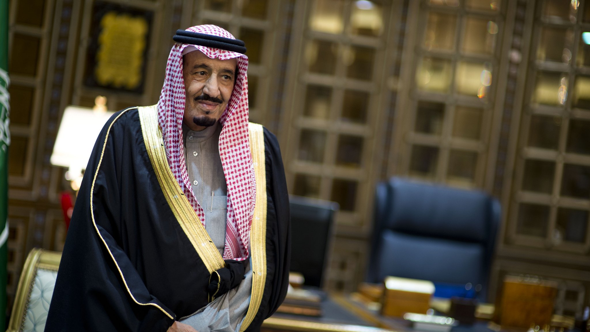 L'image montre un homme en tenue traditionnelle saoudienne, portant une robe noire ornée d'or et un keffieh, une coiffe typique. Il se tient dans un bureau richement décoré, avec des meubles en bois et un éclairage sophistiqué. L'ambiance est formelle et élégante.