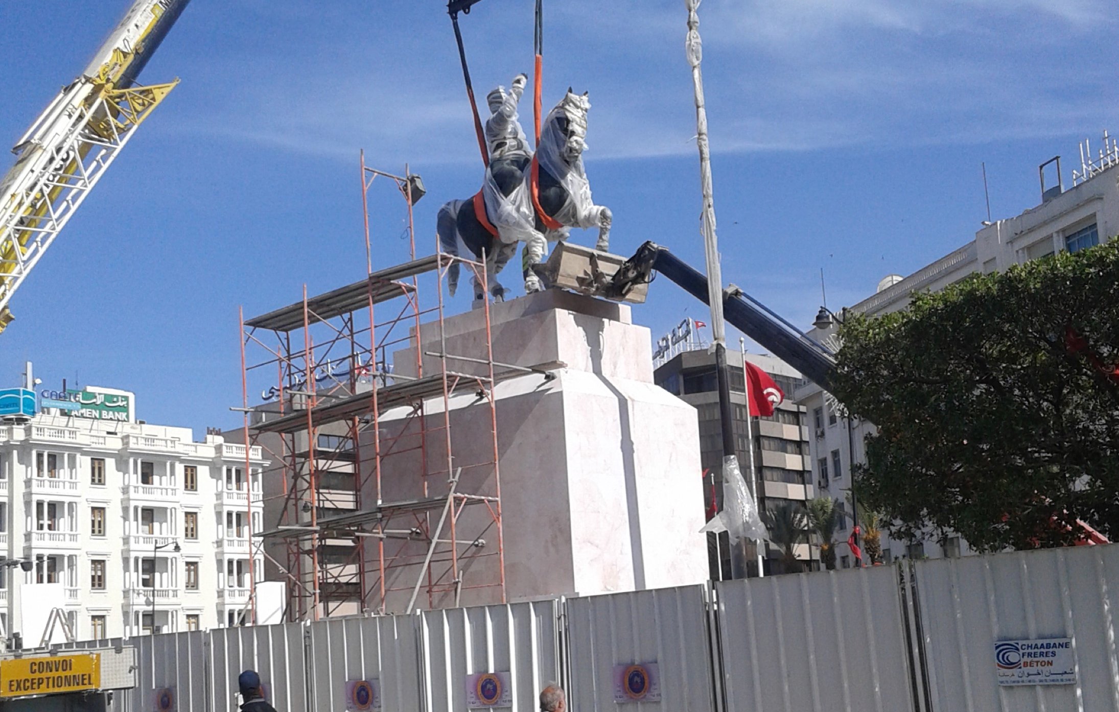 L'image montre un monument en cours d'installation. On peut voir une statue équestre accompagnée d'un cavalier, qui est en train d'être soulevée par une grue. Des échafaudages entourent la base de la statue, et un panneau de chantier est visible en arrière-plan. Le ciel est bleu, indiquant une journée ensoleillée. Des personnes marchent au premier plan, certaines portant des vêtements traditionnels.