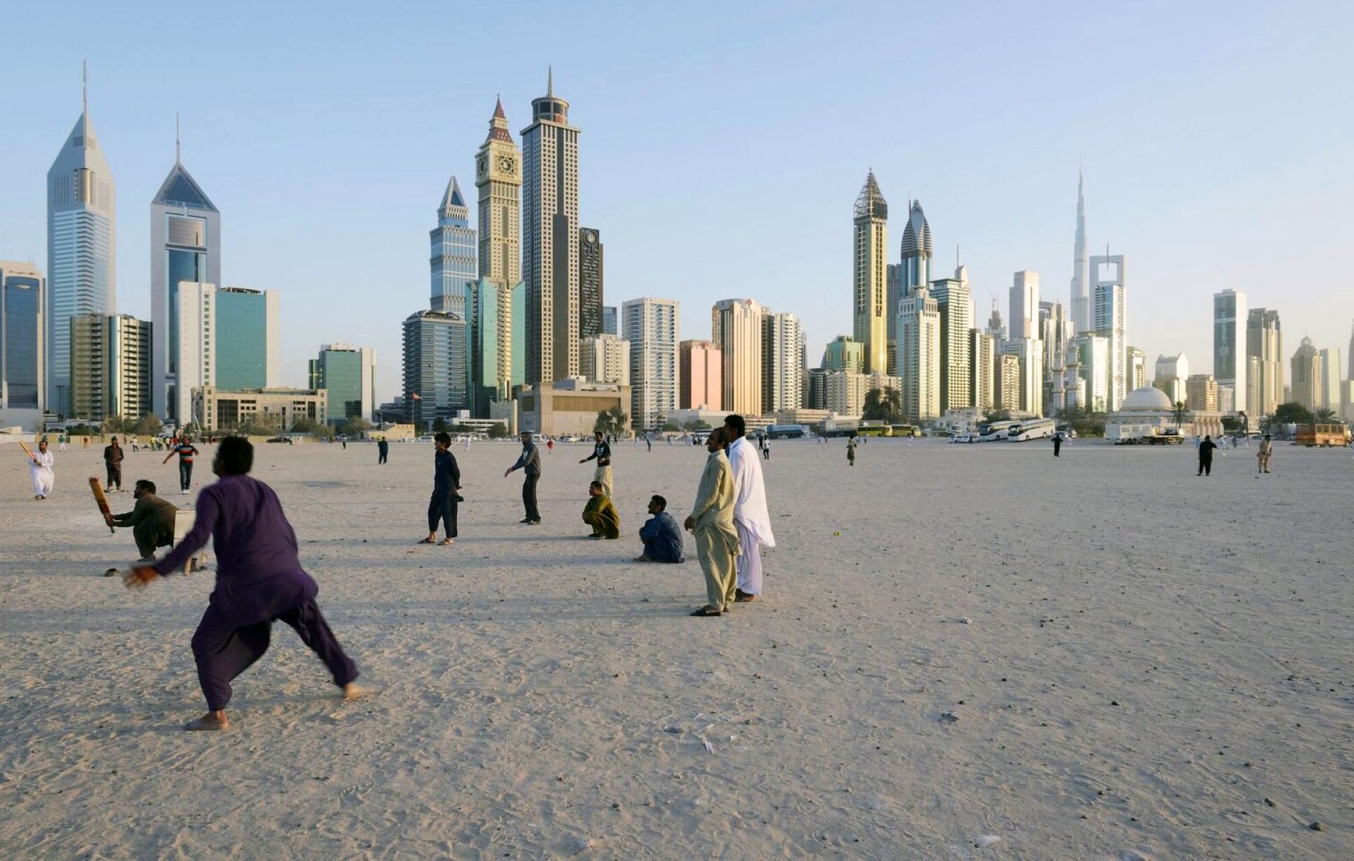 L'image montre une plage ou un espace ouvert, avec des gens qui se déplacent ou jouent dans le sable. En arrière-plan, on peut voir une ville moderne avec de grands gratte-ciel, représentant une skyline impressionnante. Les bâtiments semblent contemporains et variés en termes d'architecture. L'éclairage indique qu'il s'agit probablement d'une fin de journée, avec un ciel clair. L'atmosphère semble animée, avec des personnes profitant du lieu.