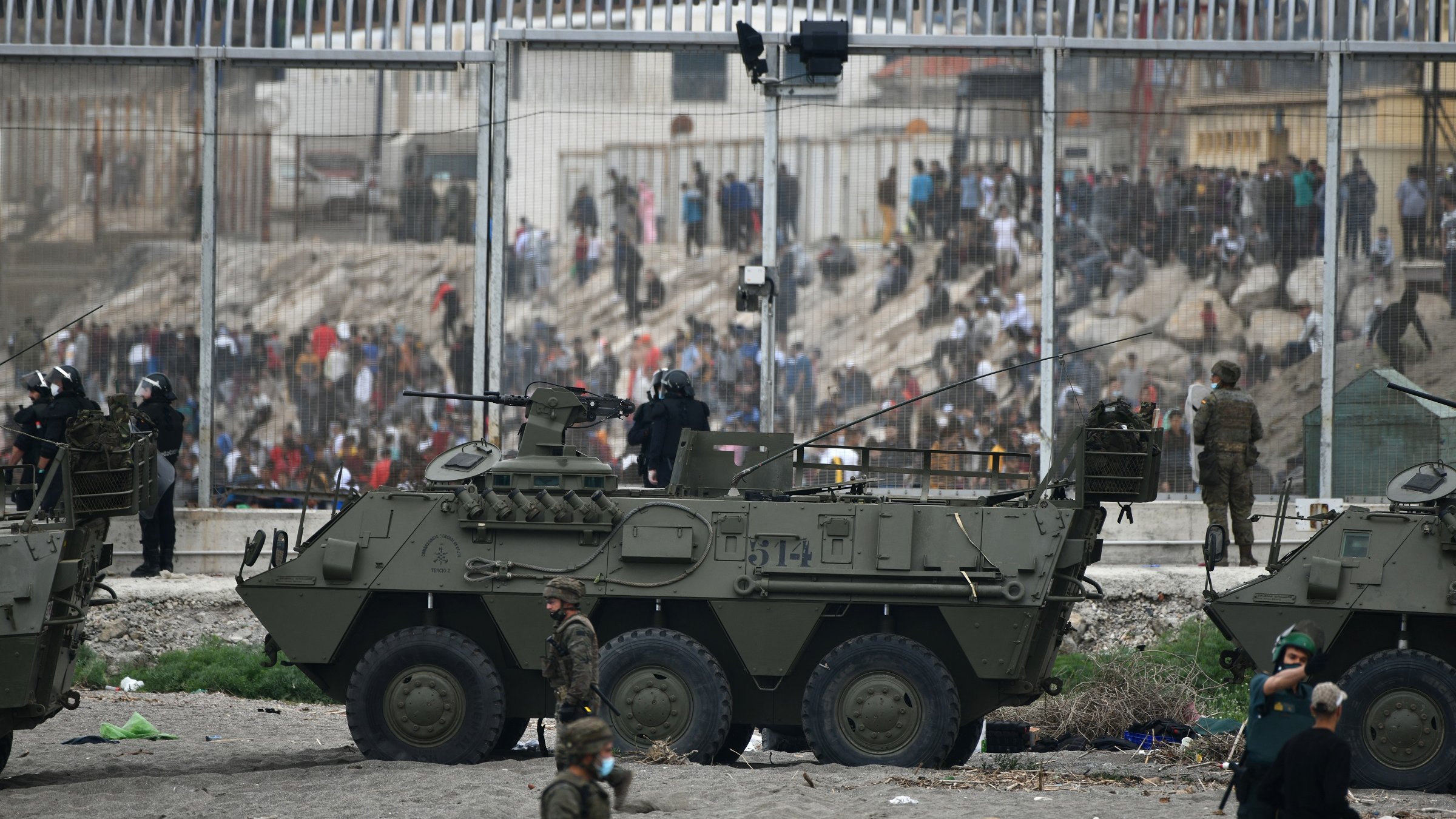 L'image montre une scène de tension, probablement dans une zone frontalière. On peut voir des véhicules militaires, dont un véhicule blindé, positionnés près d'une barrière. Des soldats sont en action, certains en uniforme et d'autres en tenue de combat, tandis qu'en arrière-plan, un grand groupe de personnes semble manifester ou se rassembler. La scène est marquée par une atmosphère de confrontation, avec des mesures de sécurité visibles.