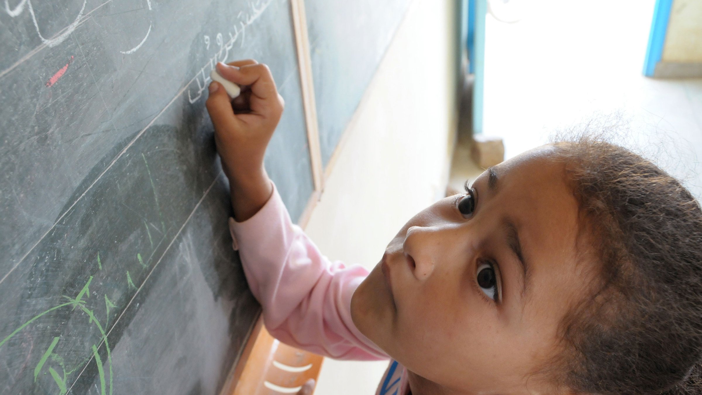 L'image montre une jeune fille penchée vers un tableau noir. Elle semble concentrée alors qu'elle écrit ou dessine quelque chose avec une craie. On peut voir des lettres ou des symboles tracés sur le tableau. La lumière entre par une fenêtre visible en arrière-plan, créant une ambiance chaleureuse. La fille porte un sweat à capuche rose et a les cheveux bouclés.