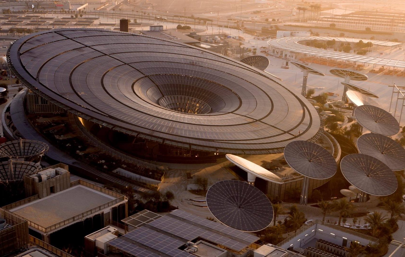 L'image montre un bâtiment architectural moderne et futuriste, avec un design en forme d'anneau ou de soucoupe volante. Le toit est en grande partie vitré, permettant à la lumière naturelle d'entrer. Des structures circulaires, semblables à des panneaux solaires ou des œuvres d'art, entourent le bâtiment, ajoutant à son aspect innovant. Le paysage environnant semble désertique, avec des infrastructures modernes visibles à l'arrière-plan, indiquant un environnement urbain en développement. La lumière douce du coucher du soleil donne une ambiance chaleureuse à la scène.