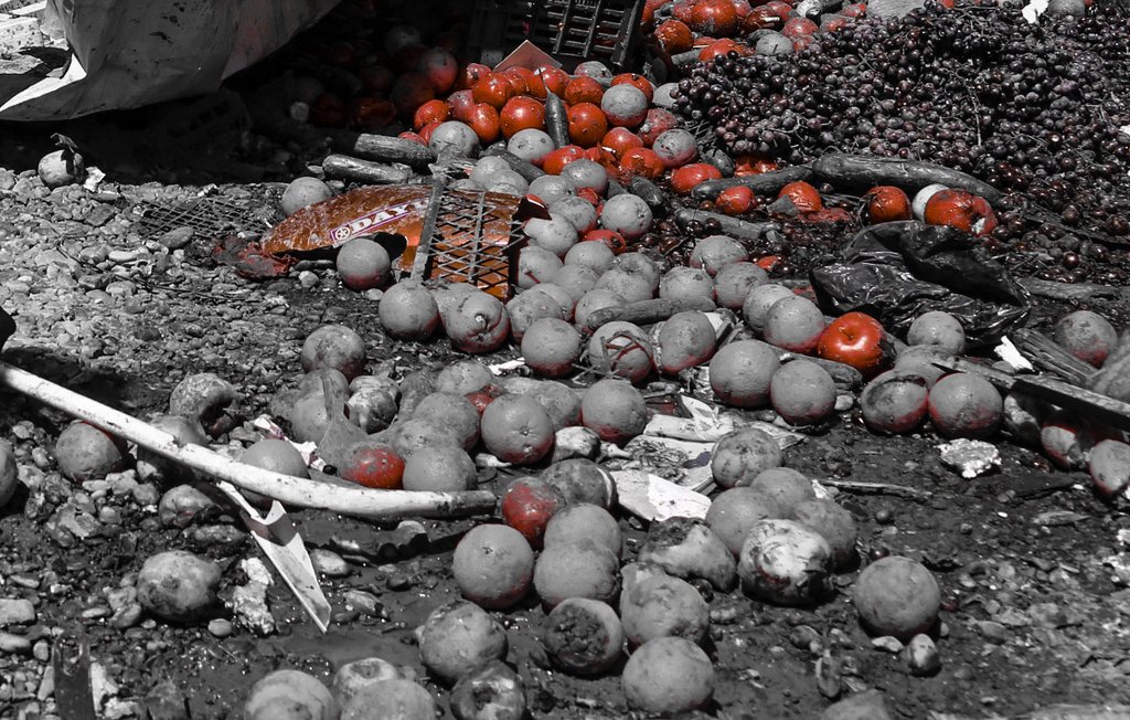 L'image montre un tas de fruits, notamment des oranges et des tomates, éparpillés sur un sol. On peut également voir des caisses en bois, certaines sont renversées, ajoutant à l'aspect chaotique de la scène. La majorité de l'image semble en noir et blanc, mais les fruits, en particulier les tomates, apparaissent en couleur, ce qui attire l'œil. Cela donne une sensation de désordre et de négligence dans ce qui semble être un marché ou un lieu de vente de fruits.