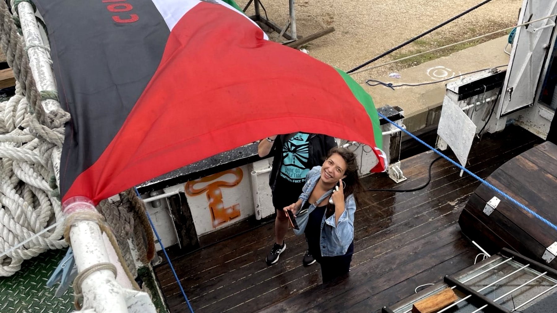 L'image montre deux personnes sur un bateau, sous un grand drapeau noir, rouge et vert. L'une des personnes sourit et pose pour la photo, tandis que l'autre semble tenir le drapeau. Le décor autour inclut du bois, des cordages et d'autres éléments d'un environnement nautique. L'atmosphère semble conviviale et dynamique.