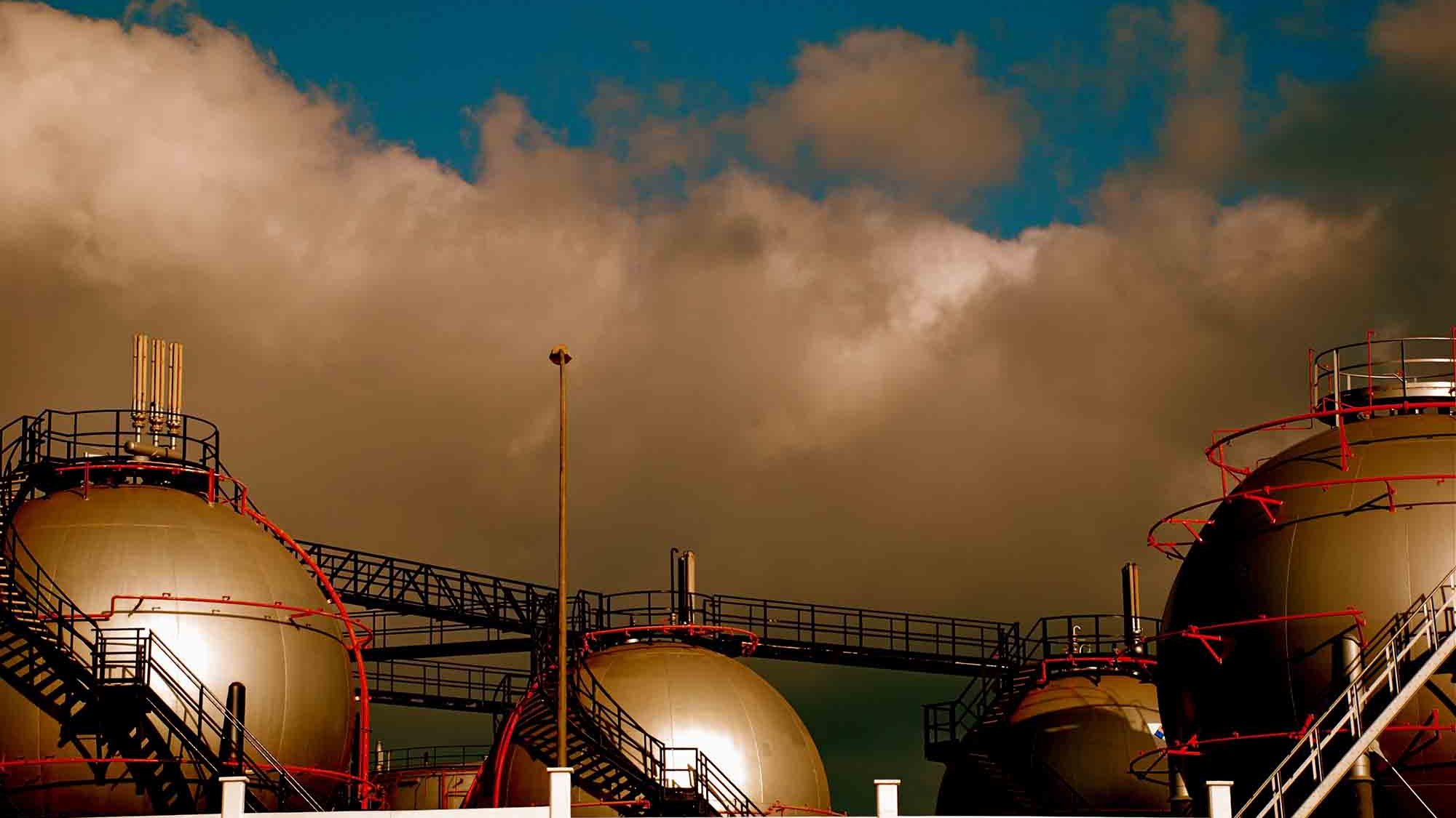 L'image présente des réservoirs à gaz de forme sphérique, surmontés de structures métalliques. Ils sont disposés sur une plateforme, tandis qu'un ciel nuageux et dramatique se profile en arrière-plan. Les couleurs à la fois sombres et lumineuses créent une atmosphère industrielle marquée par le contraste entre les réservoirs et le ciel.