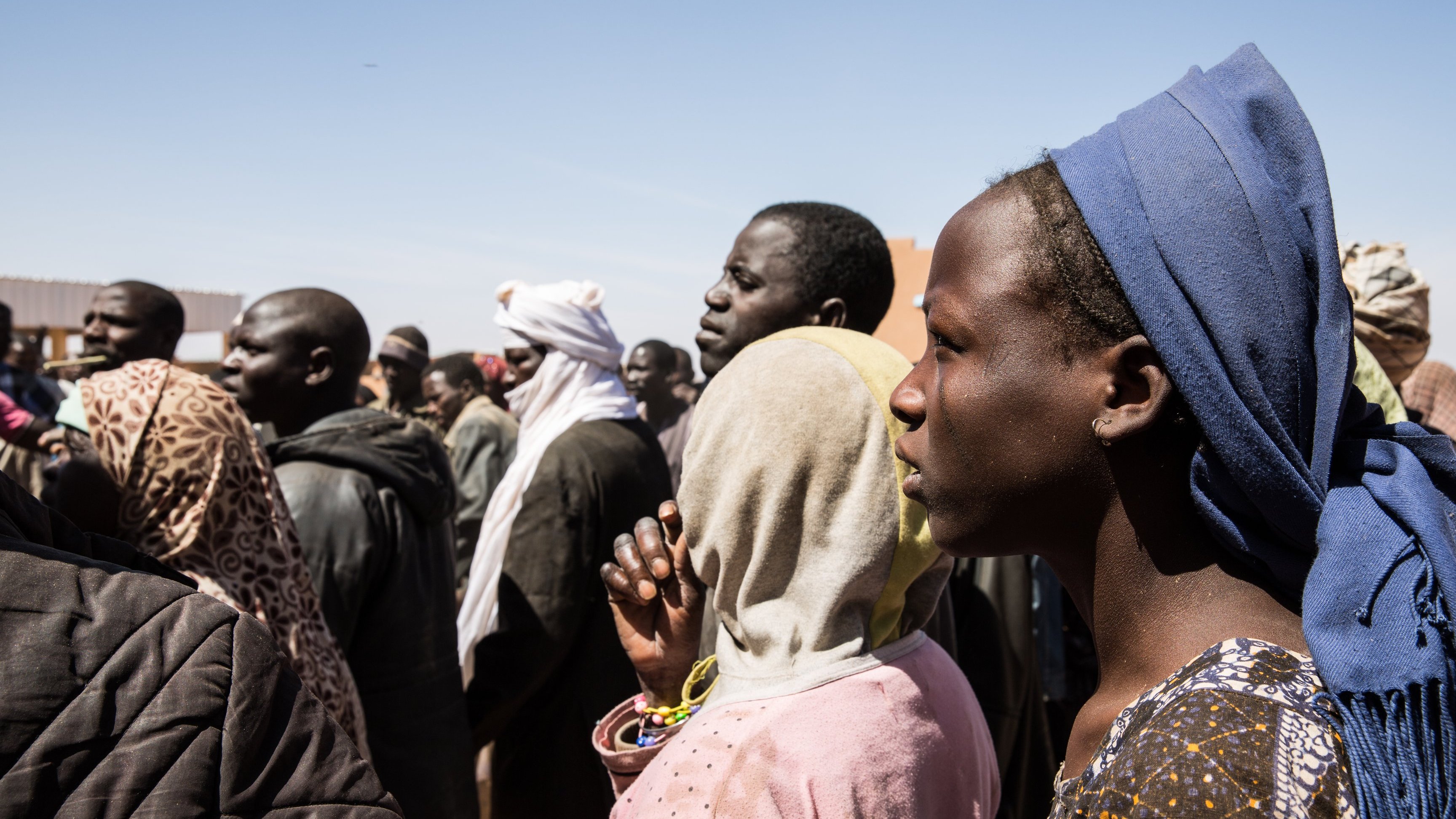 L'image montre un groupe de personnes, probablement en attente ou rassemblées pour une raison quelconque. On peut voir des visages divers avec des expressions sérieuses. Certaines personnes portent des vêtements traditionnels et des accessoires variés, comme des écharpes ou des foulards. L'arrière-plan est flou, mais le ciel est dégagé. L'ambiance semble concentrée, peut-être en lien avec un événement important.