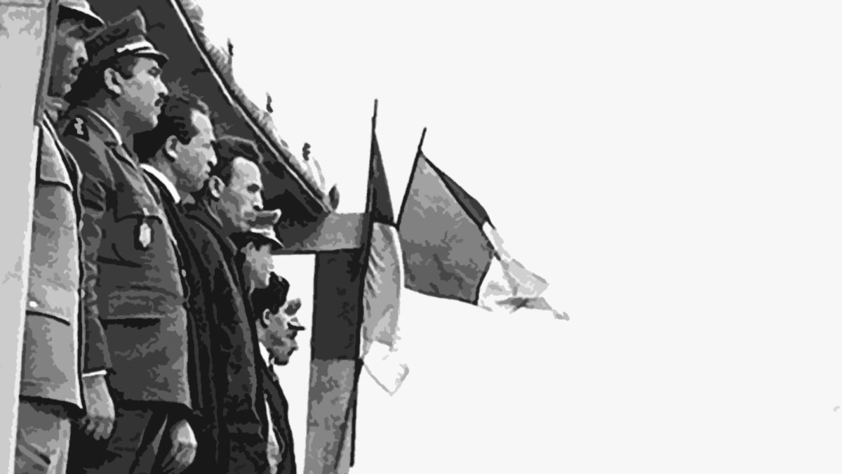 L'image montre un groupe de militaires et de dignitaires, probablement lors d'une cérémonie officielle. Ils se tiennent sur une estrade, certains en uniforme avec des médailles, tandis que d'autres portent des vêtements civils. En arrière-plan, on peut voir des drapeaux, dont un drapeau français, indiquant un contexte lié à la France ou un événement officiel. Les soldats se tiennent en position de parade, face à l'estrade. L'ambiance semble solennelle.