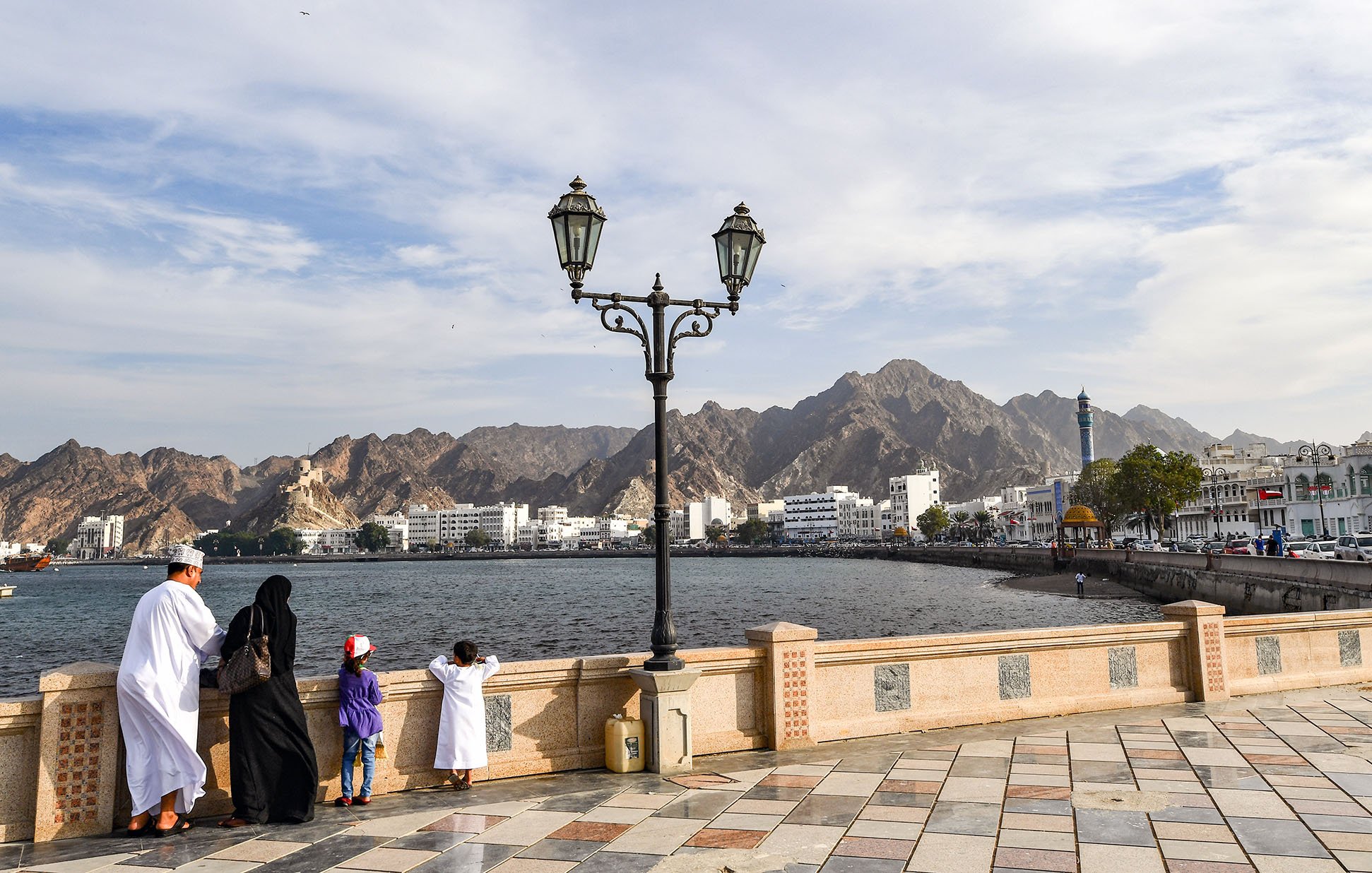 L'image montre une scène de bord de mer, probablement à Muscat, Oman. Dans le premier plan, on peut voir une famille vêtue de vêtements traditionnels. Un homme et une femme se tiennent près d'un lampadaire, regardant vers l'horizon. À côté d'eux, deux enfants, dont un porte une casquette rouge. En arrière-plan, on aperçoit des montagnes majestueuses ainsi que des bâtiments blancs typiques de la région. Le ciel est partiellement nuageux, créant une ambiance paisible et contemplative.
