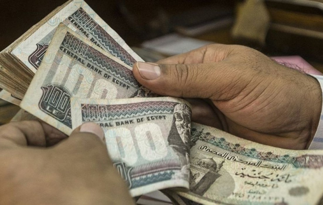 L'image montre des mains tenant des billets égyptiens. On peut voir des coupures de différentes valeurs, principalement de 100 livres égyptiennes. Les billets sont colorés et comportent des motifs et des inscriptions en arabe. L'arrière-plan semble flou, indiquant que l'attention est centrée sur l'argent.