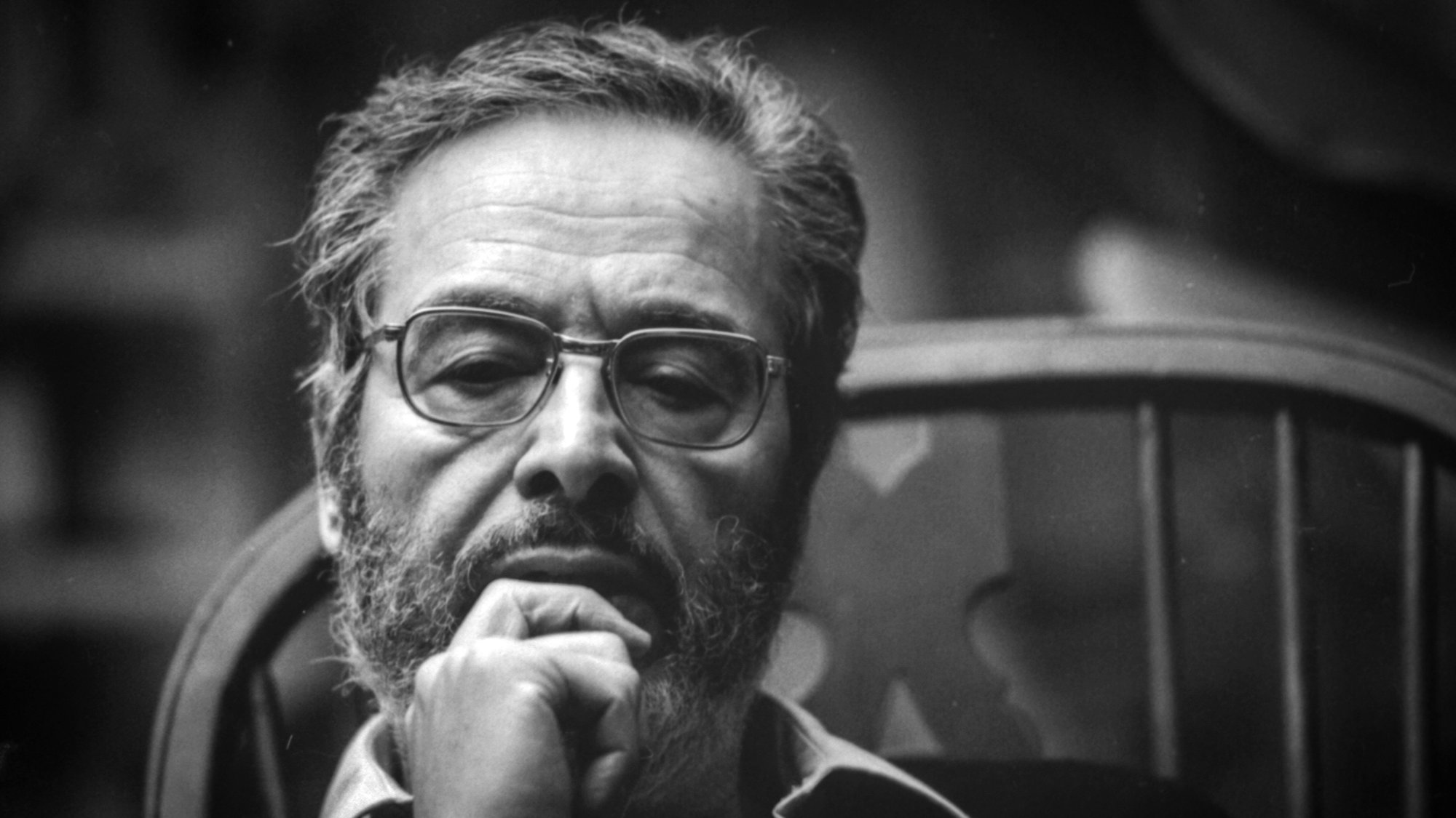 L'image montre un homme assis sur une chaise, arborant une barbe et des lunettes. Il semble profond dans ses pensées, avec une main sous le menton. L'éclairage donne un aspect sérieux à son expression, et le fond flou met en avant son visage. L'atmosphère est introspective et contemplative.