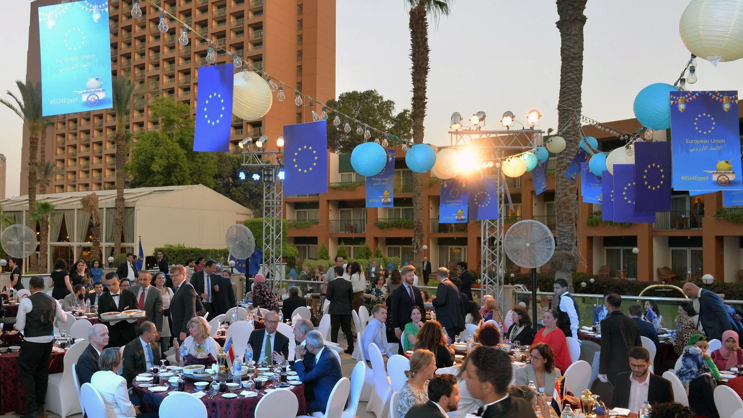 L'image montre un événement extérieur festif, probablement un dîner ou une réception. Des tables rondes sont disposées avec des nappes élégantes, où des convives en tenues formelles s'assoient pour profiter du repas. L'environnement est décoré avec des lanternes et des bannières aux couleurs de l'Union européenne. Le décor comprend également des palmiers et des installations lumineuses, créant une ambiance conviviale et festive au crépuscule. Les personnes présentes semblent converser joyeusement et profiter de l'événement.