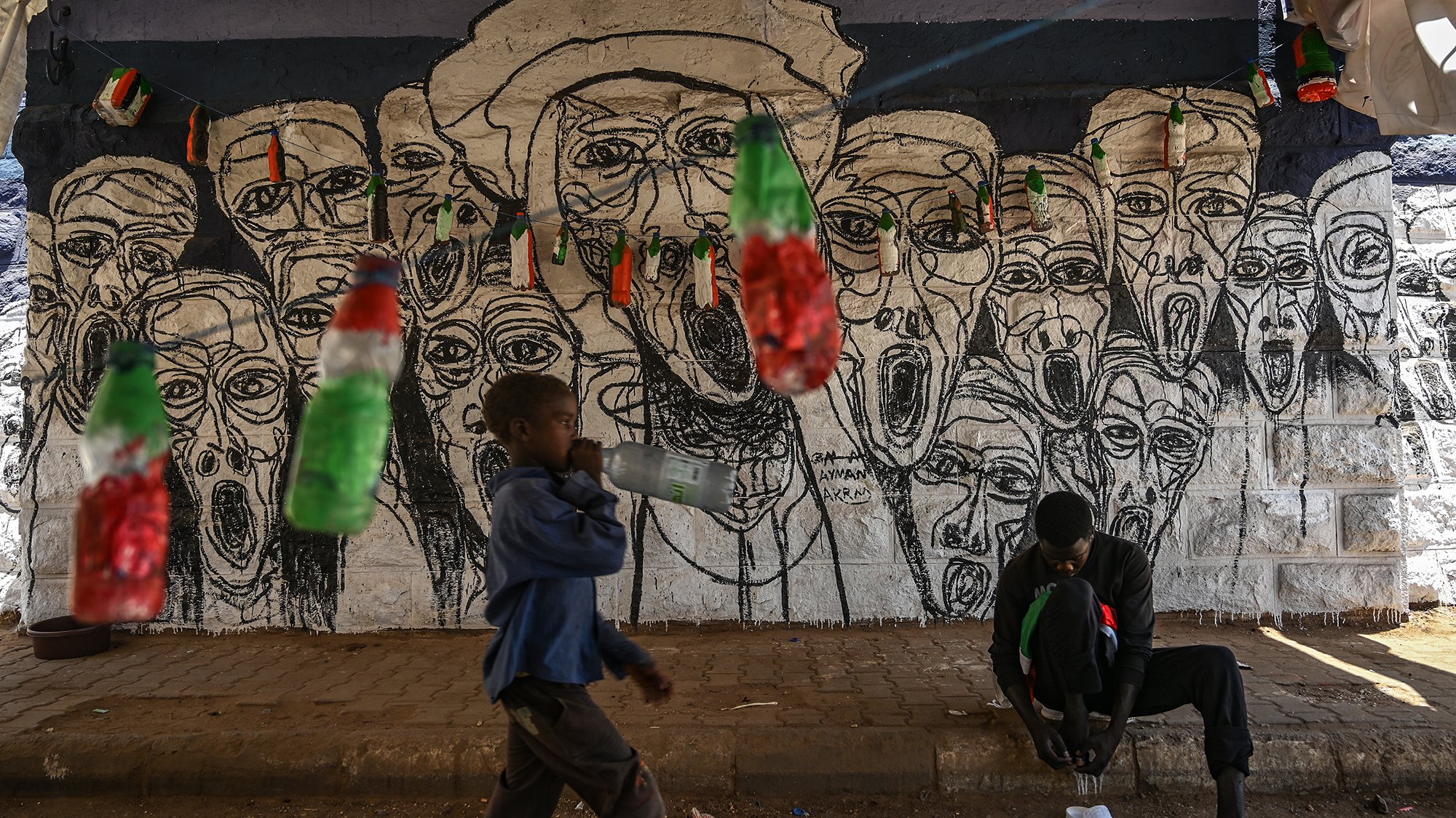 L'image montre un mur peint en arrière-plan, où se trouvent des visages aux traits exagérés, tous avec des expressions de cri, créant une atmosphère vibrante et expressive. Devant ce mur, un enfant marche en tenant une bouteille, tandis qu'un autre individu est assis au sol, occupé à lacer ses chaussures. Des bouteilles colorées, probablement en plastique, sont suspendues au-dessus d'eux, ajoutant une touche de couleur à la scène. Cette image reflète un environnement urbain dynamique, mêlant art de rue et vie quotidienne.