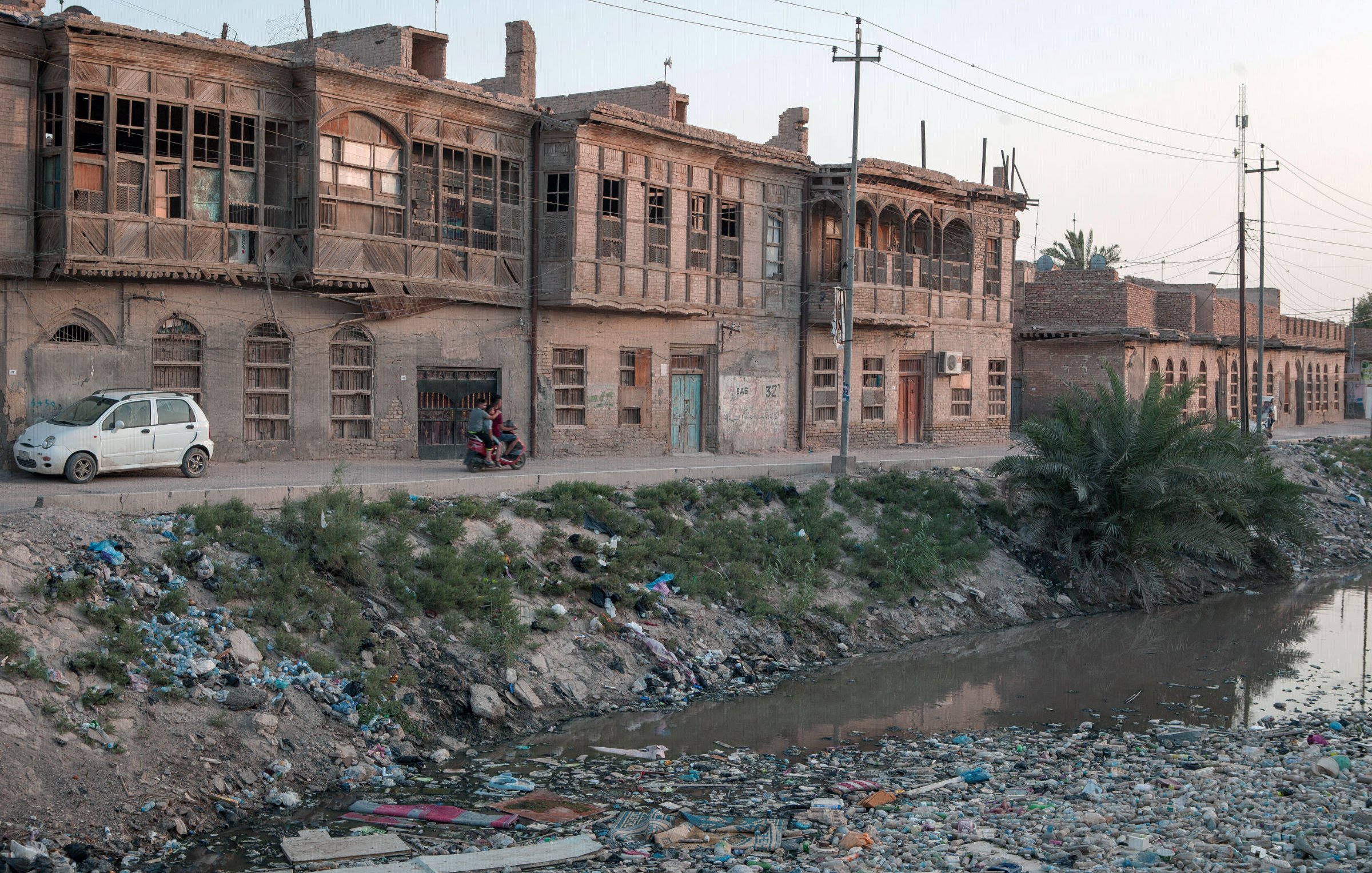 L'image montre une rue bordée de vieilles maisons en briques, caractérisées par des fenêtres en bois. À gauche, on aperçoit une voiture garée. Sur le côté droit, un groupe de personnes semble discuter. Au premier plan, un bras de rivière est visible, rempli de déchets et de débris, entouré de végétation. L'éclairage doux de la fin de journée crée une atmosphère nostalgique dans ce paysage urbain en dégradation.