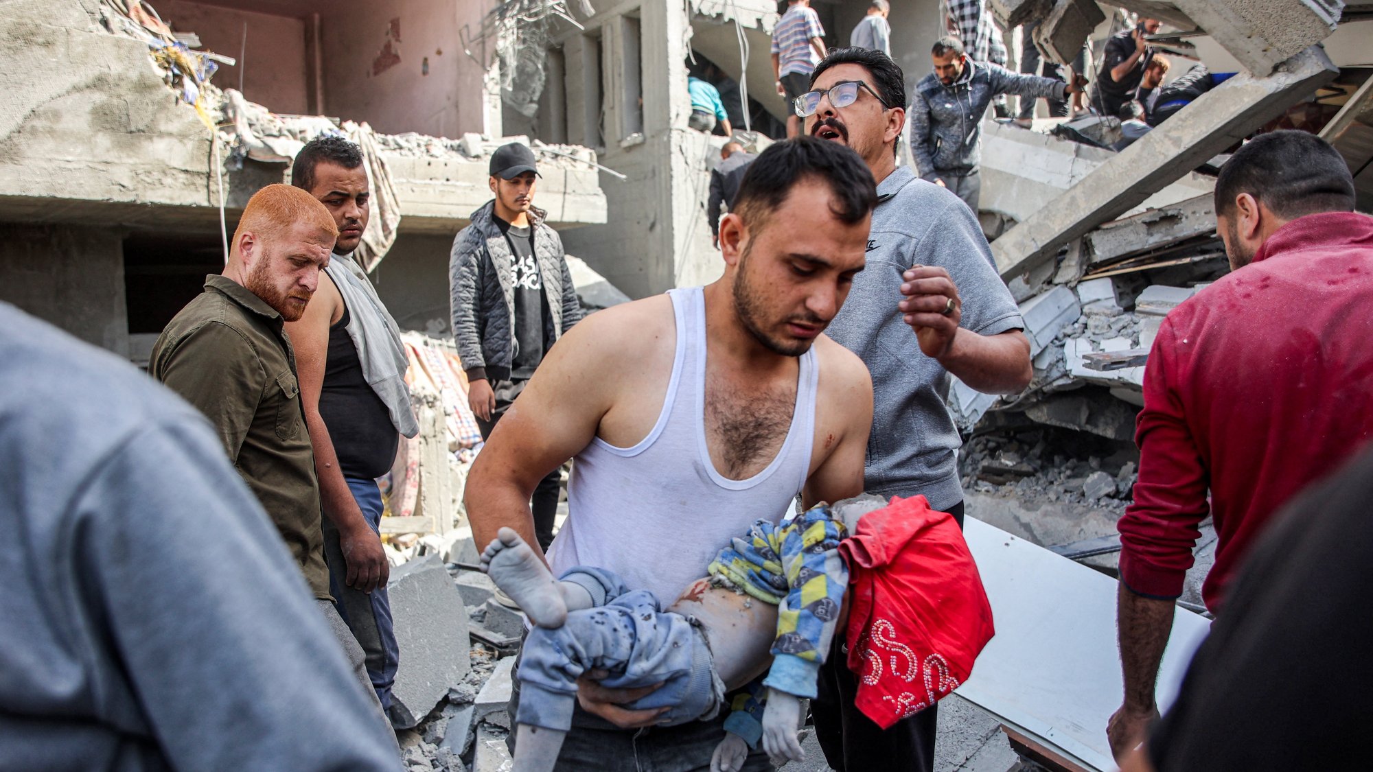 L'image montre une scène de désastre, probablement suite à un bombardement ou un tremblement de terre. On voit des décombres partout, avec des bâtiments endommagés. Un homme porte un enfant, probablement blessé, et semble préoccupé. D'autres personnes autour semblent s'affairer, cherchant à aider ou à évacuer les victimes. L'atmosphère est sombre et tragique, reflétant la détresse des personnes présentes.