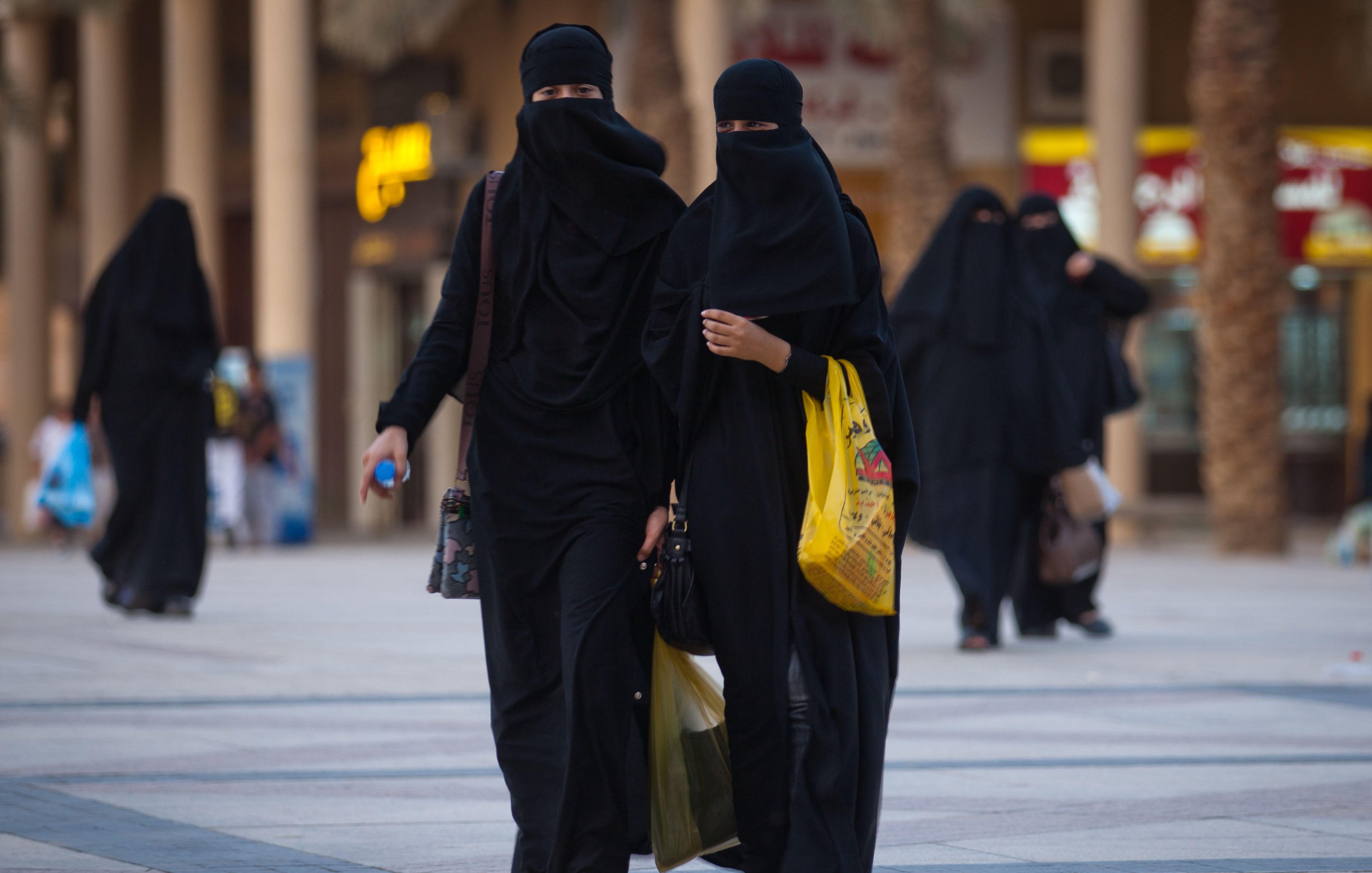 L'image montre deux femmes marchant côte à côte dans une zone urbaine. Elles portent des vêtements noirs, y compris des niqabs qui couvrent leur visage, laissant seulement les yeux visibles. L'environnement semble animé, avec des bâtiments en arrière-plan et d'autres personnes se déplaçant dans la même direction. L'une des femmes tient un sac, tandis que l'autre semble tenir un objet dans sa main. L'éclairage et l'architecture suggèrent un cadre contemporain.