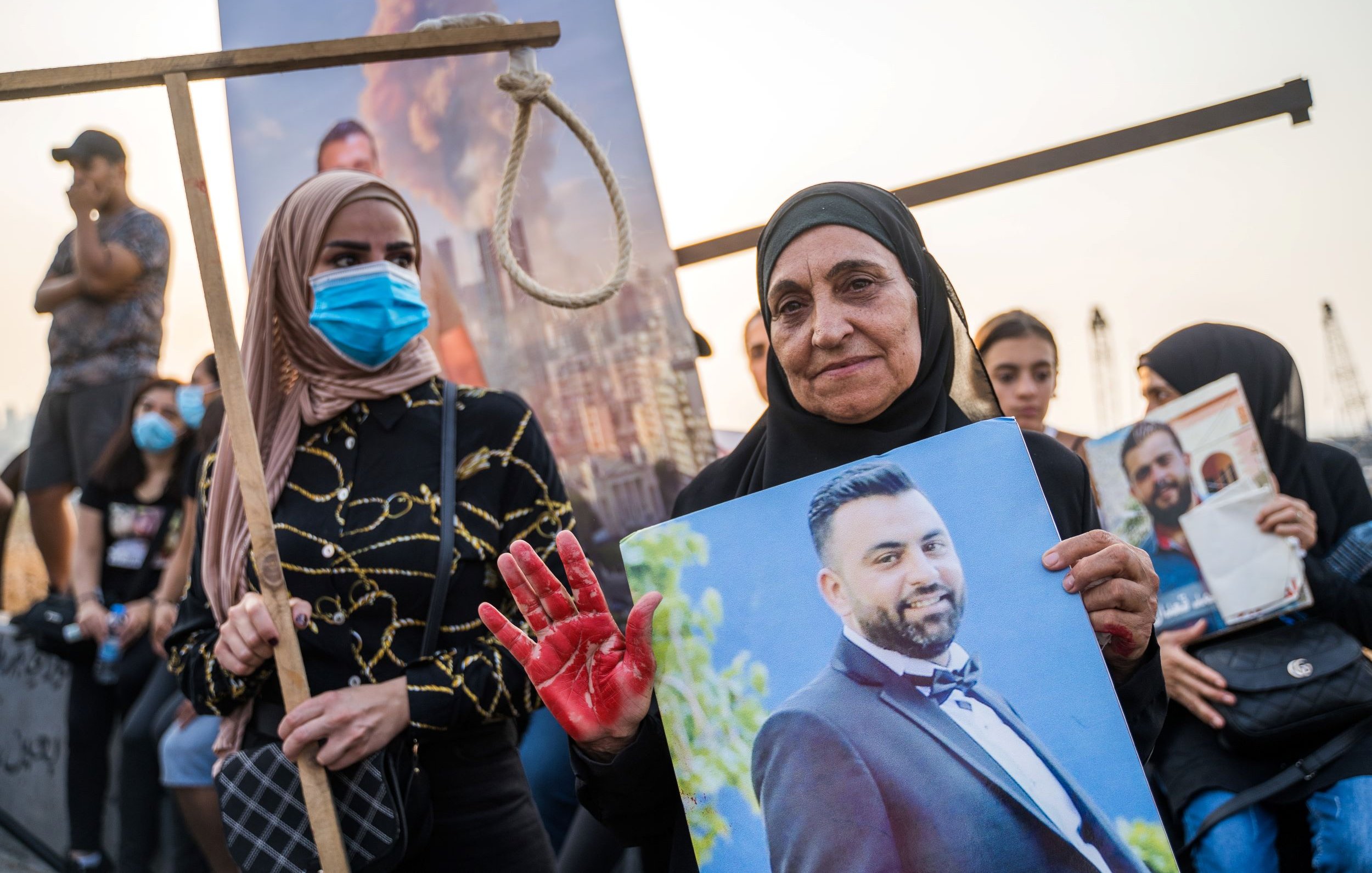 L'image montre un rassemblement commémoratif. Au premier plan, deux femmes sont présentes. L'une porte un hijab noir et tient une grande photo d'un homme. L'autre femme, à sa gauche, porte un masque bleu et semble également participer à la manifestation. La femme avec la photo a une main teintée de rouge, symbolisant peut-être la douleur ou la protestation. En arrière-plan, on peut voir des personnes rassemblées, ainsi qu'un décor urbain qui suggère un contexte de tension ou de lutte.