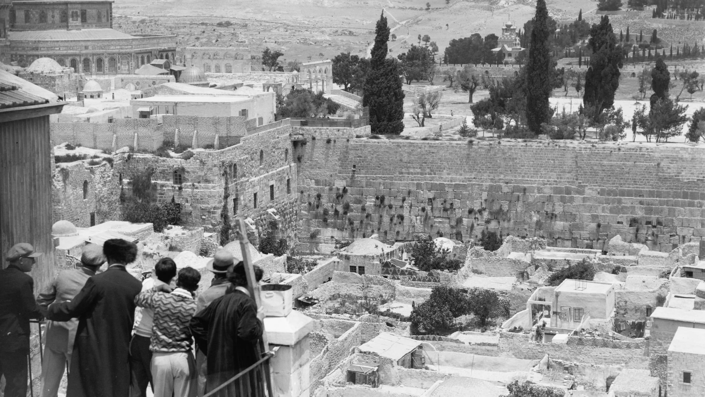 L'image montre une vue panoramique d'un paysage urbain historique. Au premier plan, un groupe de personnes se tient sur un balcon ou une terrasse, observant la scène devant eux. On aperçoit des ruines et des bâtiments anciens, tandis qu'en arrière-plan, on distingue des collines et des arbres. La célèbre mosquée du Dôme du Rocher est visible, ajoutant une dimension significative à cette scène. Le ciel est partiellement nuageux, et l'image évoque une atmosphère de calme et de contemplation.