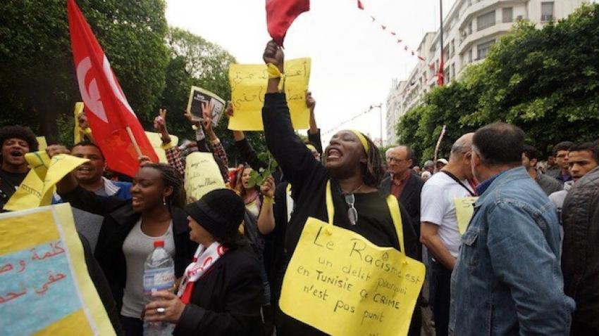 L'image montre une manifestation dans une rue animée, avec des gens qui tiennent des pancartes et des drapeaux. Une personne au centre, habillée en noir, lève un panneau sur lequel est écrit "Le Racisme est un délit et un crime en Tunisie". Les manifestants semblent exprimer des préoccupations concernant le racisme. L'ambiance est vibrant et engagée, avec des participants qui chantent ou crient des slogans. Les éléments visuels suggèrent une mobilisation sociale pour des droits et la justice.