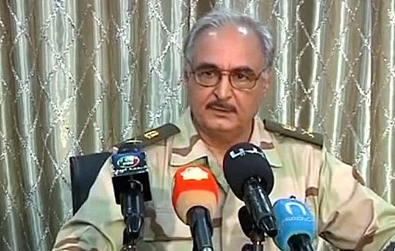 L'image montre un homme portant un uniforme militaire avec des rangs. Il est assis devant plusieurs microphones, suggérant qu'il est en train de donner une déclaration ou une interview. L'arrière-plan est composé de rideaux clairs, et l'homme a une expression sérieuse. On sent qu'il s'agit d'une situation officielle.