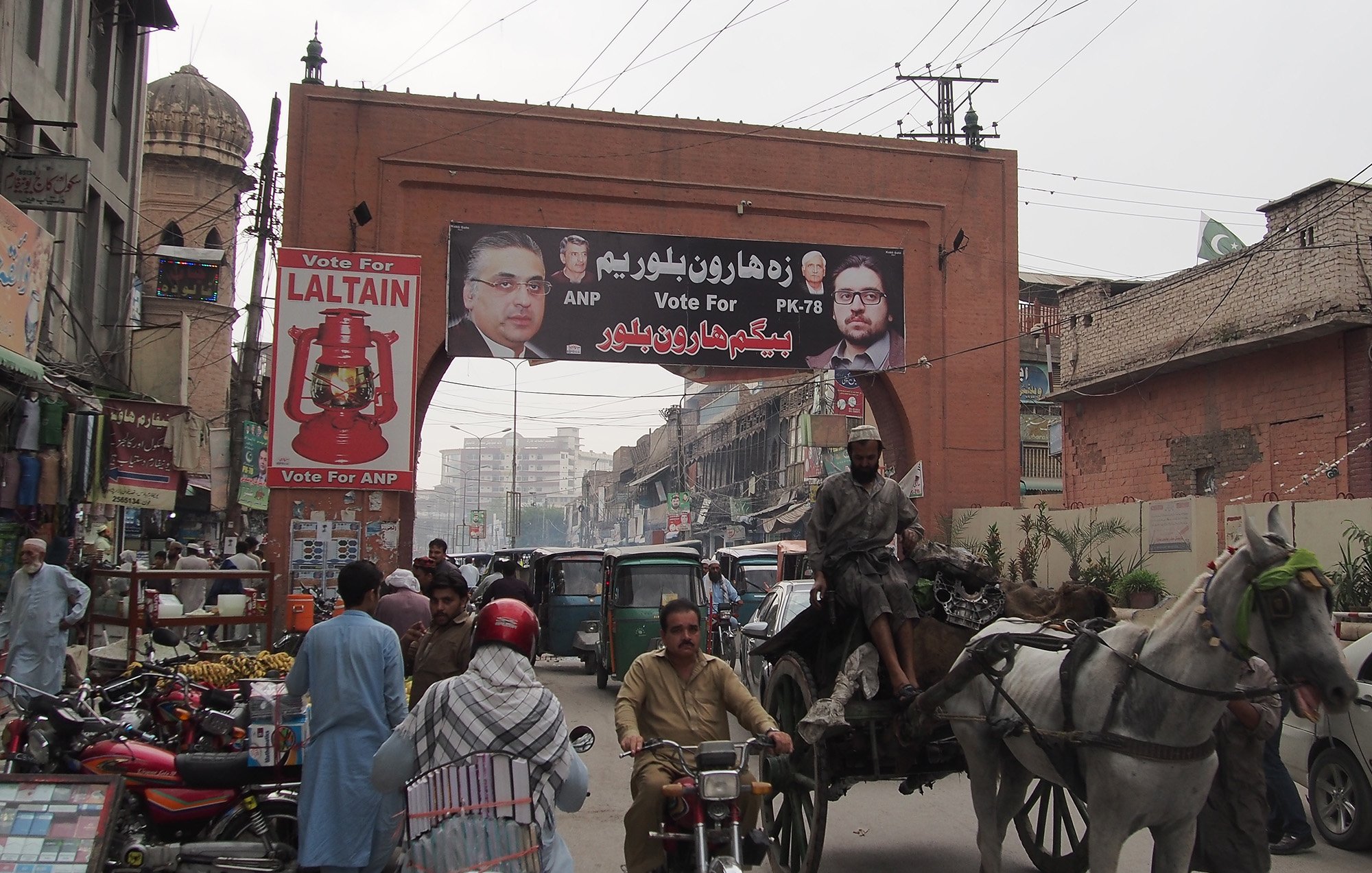 L'image montre une rue animée d'une ville avec un grand panneau publicitaire suspendu au-dessus. Ce panneau affiche des portraits de candidats politiques, accompagnés d'un message de campagne. Au premier plan, on voit des véhicules variés, y compris des charrettes tirées par des chevaux, des motos et des bicyclettes, illustrant l'activité de la vie urbaine. En arrière-plan, on peut apercevoir des bâtiments typiques et une ambiance dynamique, avec des personnes se déplaçant dans la rue.