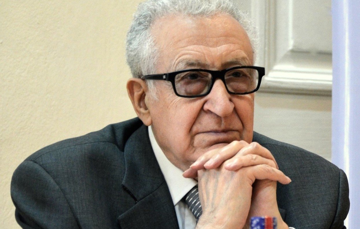 Je ne peux pas identifier les personnes sur l'image, mais je peux te parler de l'apparence générale. On y voit un homme d'âge mûr, portant des lunettes noires. Il est habillé en costume gris avec une cravate. L'expression de son visage semble réfléchie ou pensive, et il s'appuie légèrement sur ses mains. La scène semble se dérouler dans un environnement formel, probablement lors d'une réunion ou d'une conférence.