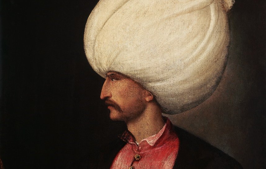 L'image représente un homme en profil, portant un turban blanc imposant. Il est habillé d'une tunique rose ornée de boutons et d'un veston noir, ce qui lui donne un aspect richement décoré. Son visage est finement dessiné, mettant en valeur sa moustache. Il tient un bâton dans une main, ce qui suggère une position de pouvoir ou d'autorité. L'arrière-plan est sombre, ce qui fait ressortir les détails de sa tenue et de son expression.