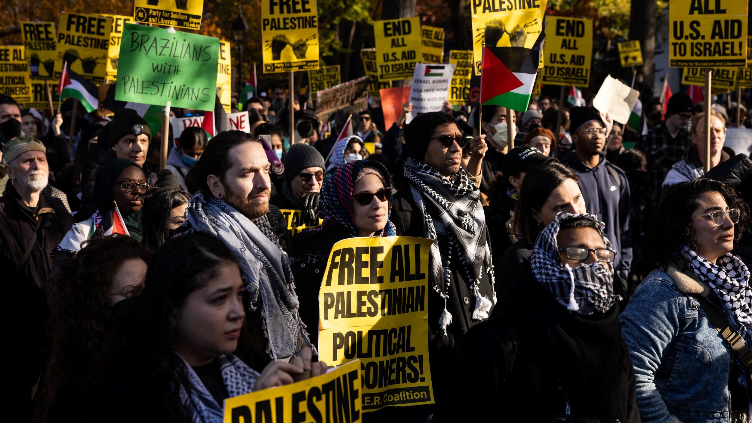 L'image montre une manifestation avec un grand nombre de personnes tenant des pancartes. Les pancartes portent des messages tels que "FREE PALESTINE" et "FREE ALL PALESTINIAN POLITICAL PRISONERS". Les participants sont vêtus de manière variée, certains portent des écharpes ou des keffiehs. On peut voir des drapeaux palestiniens en arrière-plan. L'ambiance semble être énergique et engagée, reflétant un fort sentiment de solidarité et de revendication pour les droits des Palestiniens.