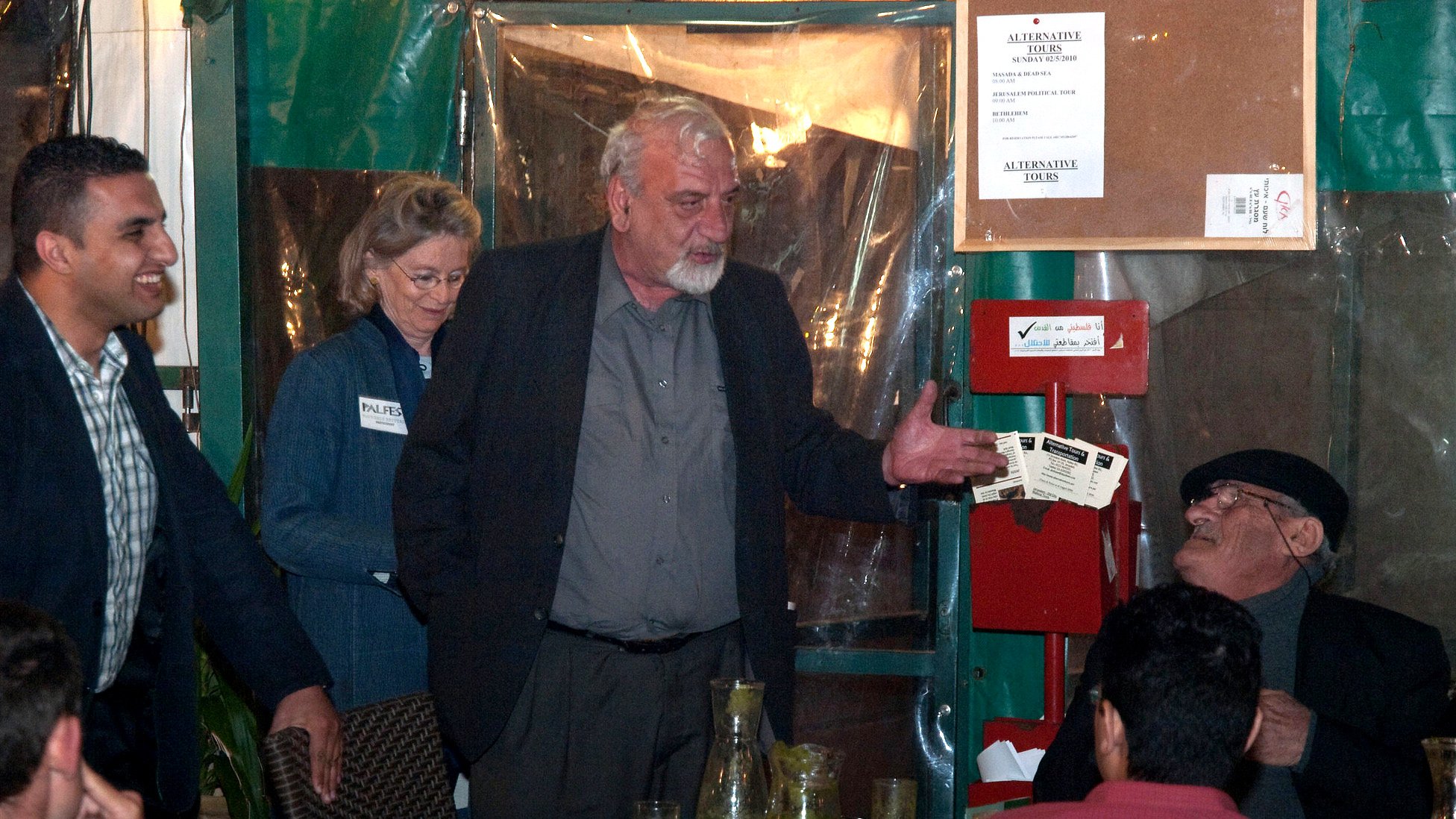 L'image montre un groupe de personnes rassemblées dans un espace qui semble être un restaurant ou une salle de réunion. Un homme au centre, avec des cheveux gris et portant une chemise noire, semble être en train de parler ou de gesticuler. À sa gauche, une femme avec des cheveux ondulés est debout et regardent vers lui. Un autre homme, assis, écoute attentivement, tandis que d'autres personnes sont également présentes à la table. L'environnement est décontracté, avec des éléments typiques de la restauration, tels que des verres et des boissons sur la table.