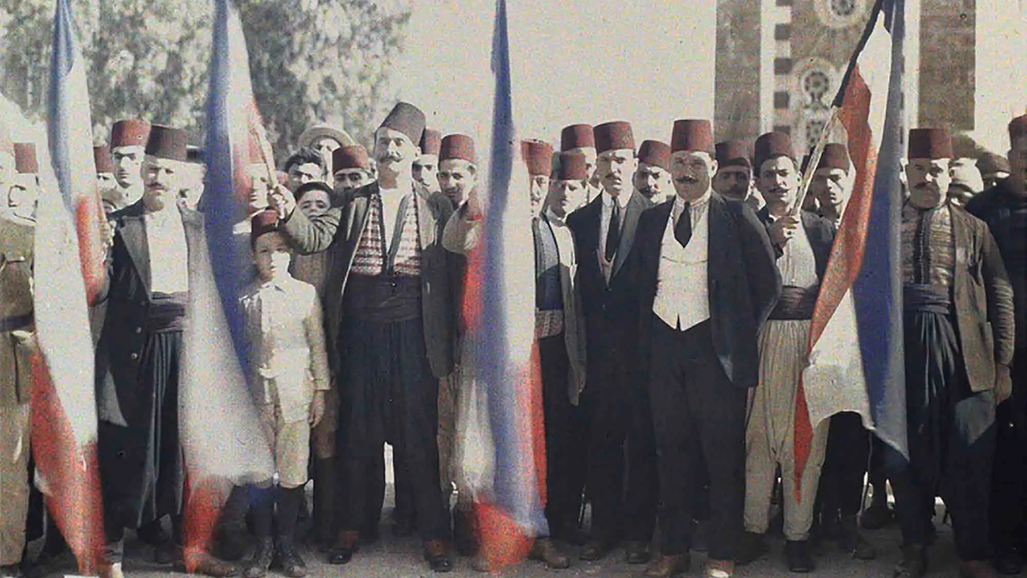L'image montre un groupe de personnes en tenue traditionnelle, se tenant ensemble avec des drapeaux russes. Les hommes portent des fez et divers vêtements folkloriques, avec un jeune garçon au centre. L'arrière-plan présente une architecture qui semble historique, peut-être un bâtiment symbolique. L'atmosphère suggère un moment de célébration ou de rassemblement associé à une certaine fierté nationale.