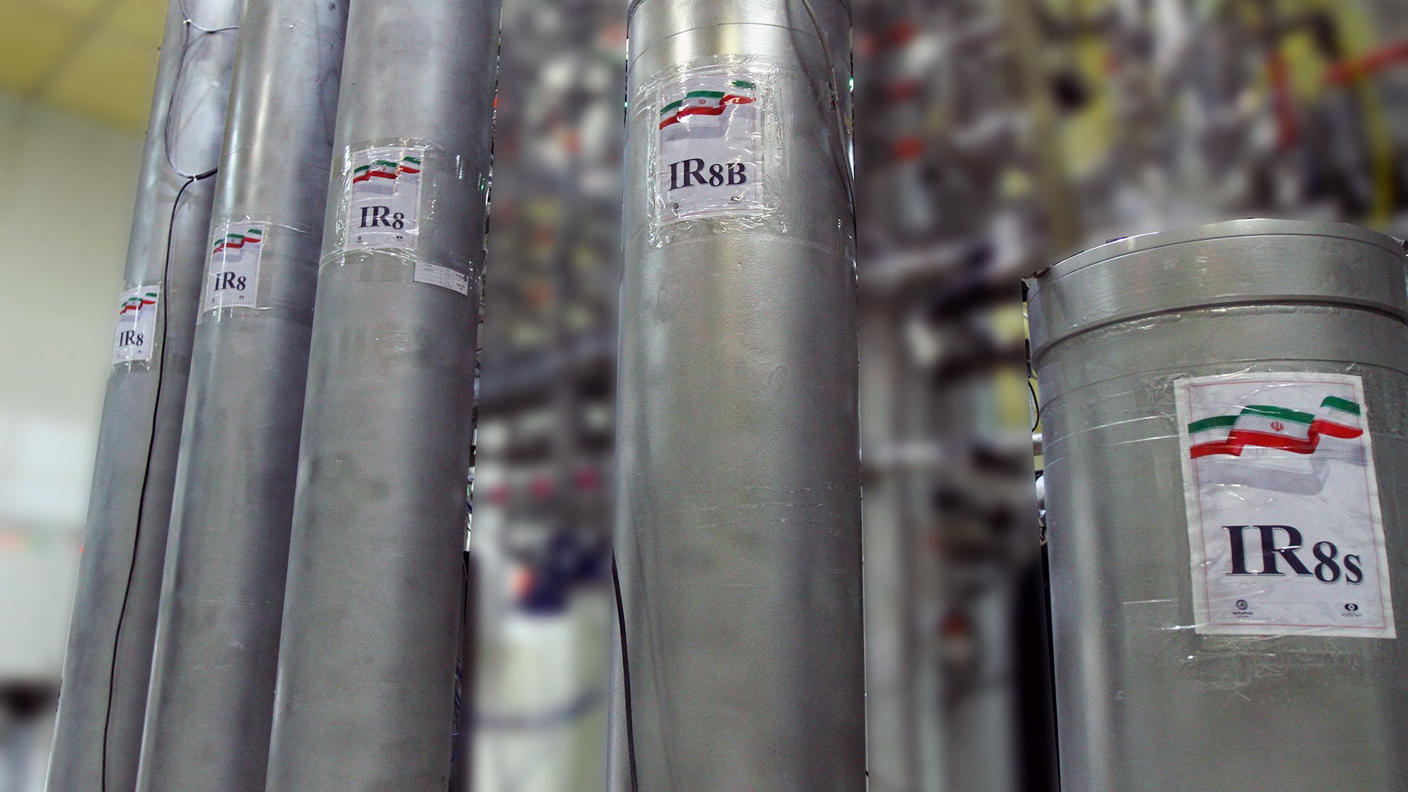 L'image montre plusieurs cylindres métalliques étiquetés avec des inscriptions visibles, y compris "IR8" et "IR8b". Ces cylindres semblent faire partie d'une installation technique, probablement liée à l'enrichissement de l'uranium, étant donné leur apparence et le contexte. En arrière-plan, on peut apercevoir d'autres équipements complexes, suggérant un environnement industriel ou scientifique avancé. Les étiquettes affichent également des drapeaux stylisés, probablement en référence à un pays spécifique.