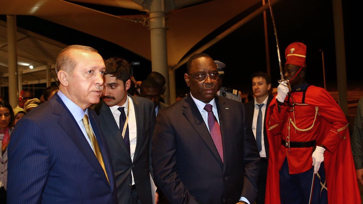 L'image montre deux hommes en costume, marchant ensemble dans un cadre officiel. L'un d'eux semble être un dirigeant turc, tandis que l'autre représente le Sénégal. Ils sont entourés de plusieurs personnes en arrière-plan, et un garde d'honneur en uniforme rouge se tient à proximité. L'atmosphère paraît sérieuse et formelle, typique d'une rencontre diplomatique.
