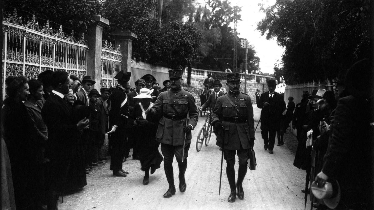 L'image montre une rue entourée de personnes, dont des militaires en uniforme. Deux officiers marchent au premier plan, semblant discuter. On aperçoit des civils sur les côtés, certains regardant vers les officiers. L'ambiance semble solennelle, et l'environnement est arboré, avec des bâtiments en arrière-plan. Les vêtements des personnes indiquent une époque historique. L'image a une qualité noire et blanche, typique des photographies anciennes.