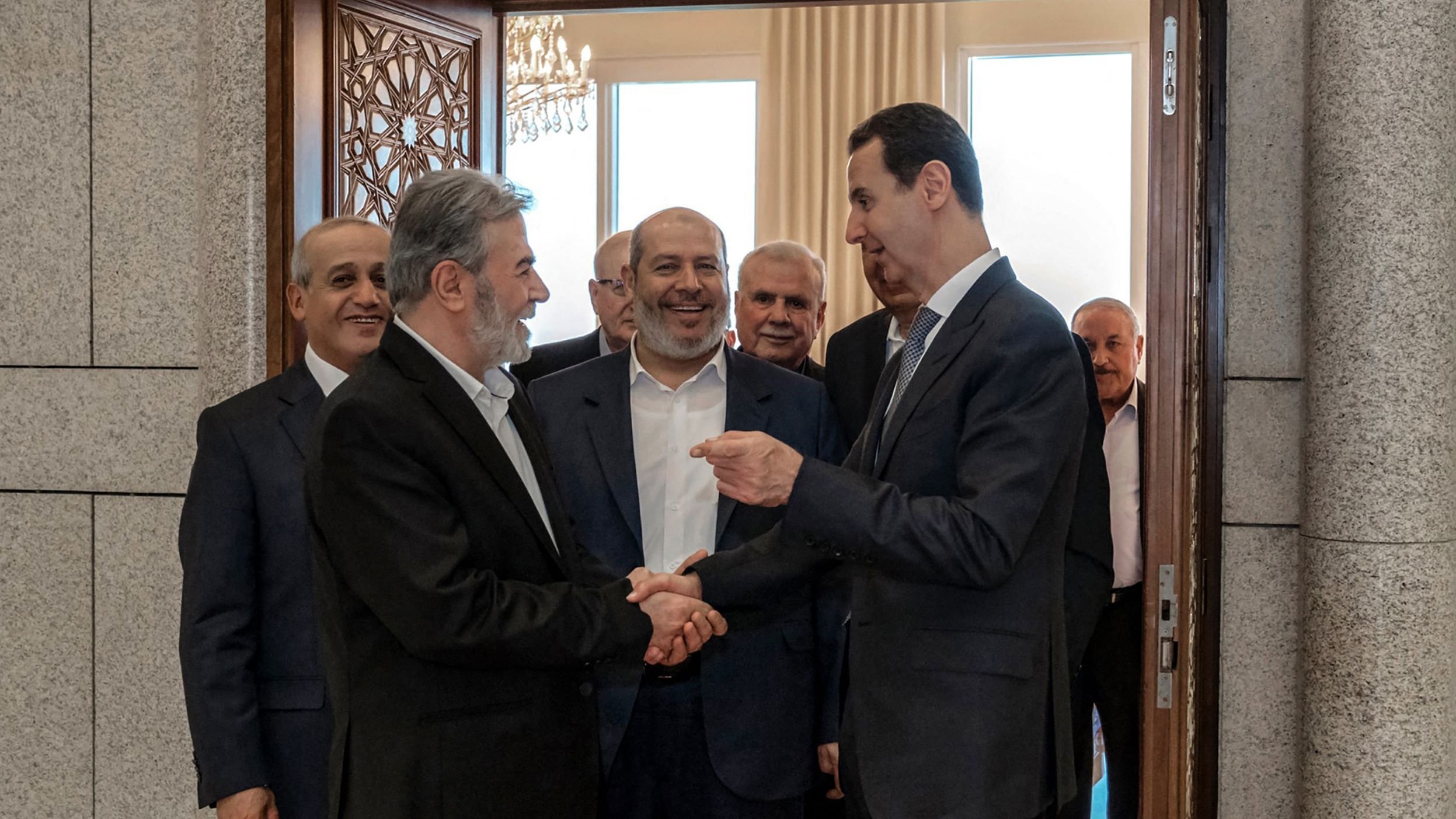 L'image montre un groupe d'hommes en train de se rencontrer dans un intérieur. Plusieurs d'entre eux sourient et semblent engagés dans une conversation amicale. L'un des hommes, au premier plan, serre la main d'un autre, tandis que les autres les observent. L'environnement semble formel, avec des éléments décoratifs visibles, comme une porte ornée et des fenêtres laissant entrer la lumière.