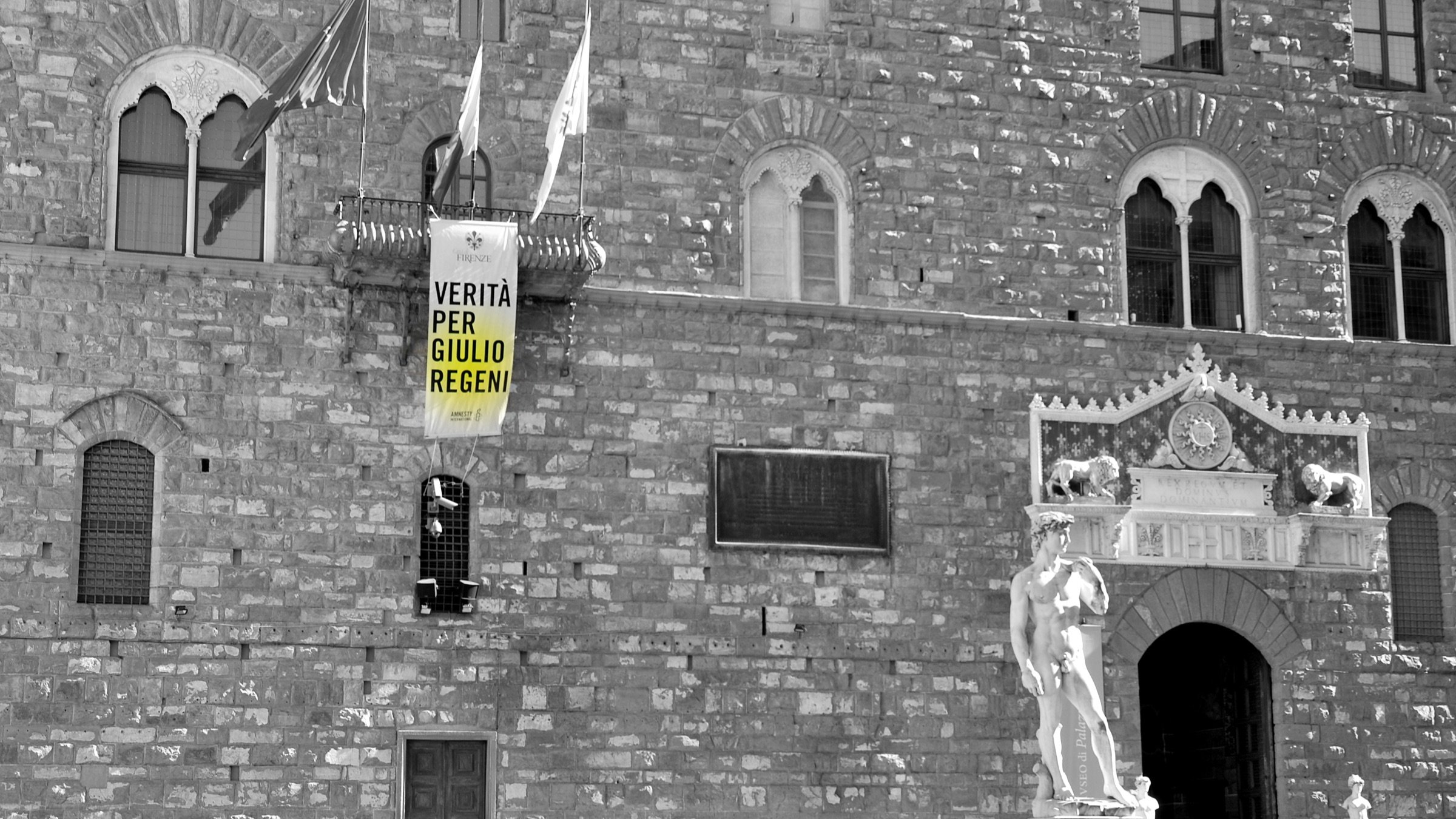 L'image montre un bâtiment historique avec une façade en pierre. Il y a une grande banderole accrochée à la façade, sur laquelle on peut lire "VERITÀ PER GIULIO REGENI", ce qui signifie "Vérité pour Giulio Regeni". En bas, on aperçoit une statue blanche, sûrement une œuvre d'art classique. Plusieurs personnes se déplacent devant le bâtiment, créant une ambiance animée. L'image est en noir et blanc, ce qui accentue le sérieux du message sur la banderole.