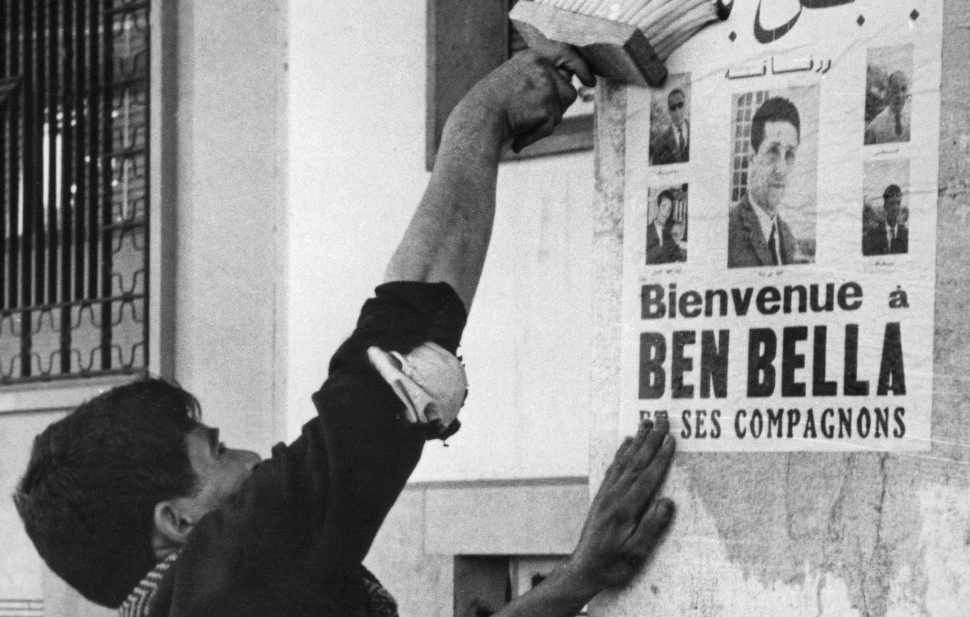L'image montre un homme en train de coller une affiche sur un mur. L'affiche annonce un message de bienvenue à Ben Bella et ses compagnons, tout en affichant plusieurs portraits. L'homme semble concentré et utilise un outil pour bien lisser l'affiche contre la surface. L'environnement urbain suggère une période historique, probablement liée à un événement important. Les détails architecturaux et la tenue de l'homme donnent une impression de contexte social et culturel spécifique.