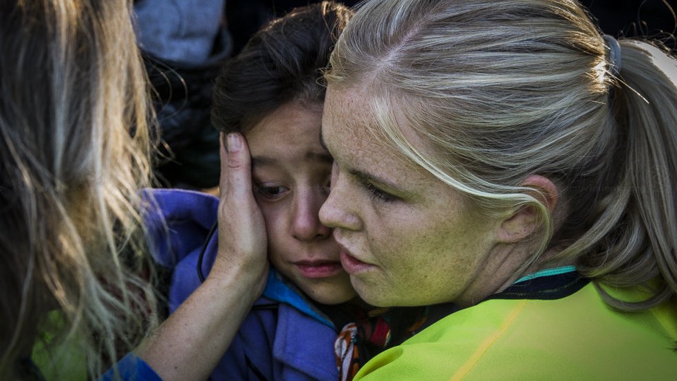 L'image montre une scène émotive où une femme, vêtue de vêtements de couleur vive, réconforte une petite fille. La femme semble douce et protectrice, tenant délicatement le visage de l'enfant. La petite fille a l'air triste ou inquiète, et elle se tourne vers la femme pour chercher du réconfort. L'environnement semble être chargé d'émotion, avec des personnes présentes en arrière-plan. Cette interaction illustre un moment de soutien et de compassion.