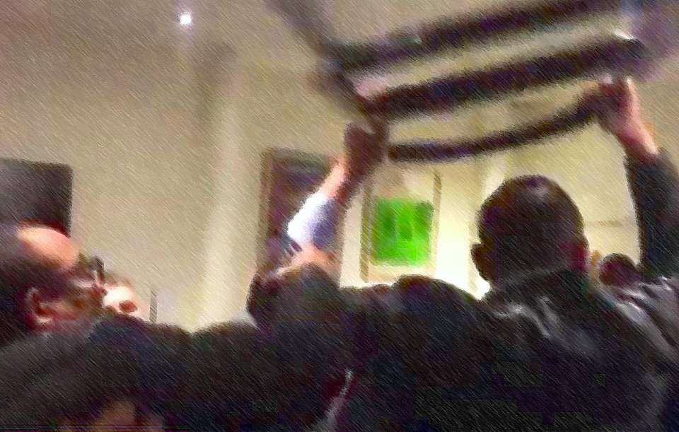 L'image montre un groupe de personnes se rassemblant autour d'une bannière ou d'un drapeau, qu'ils soulèvent. L'atmosphère semble dynamique et engagée, avec des individus exprimant leurs émotions. Le fond est flou, ce qui met l'accent sur l'action au premier plan. On peut sentir une ambiance de solidarité ou de protestation.