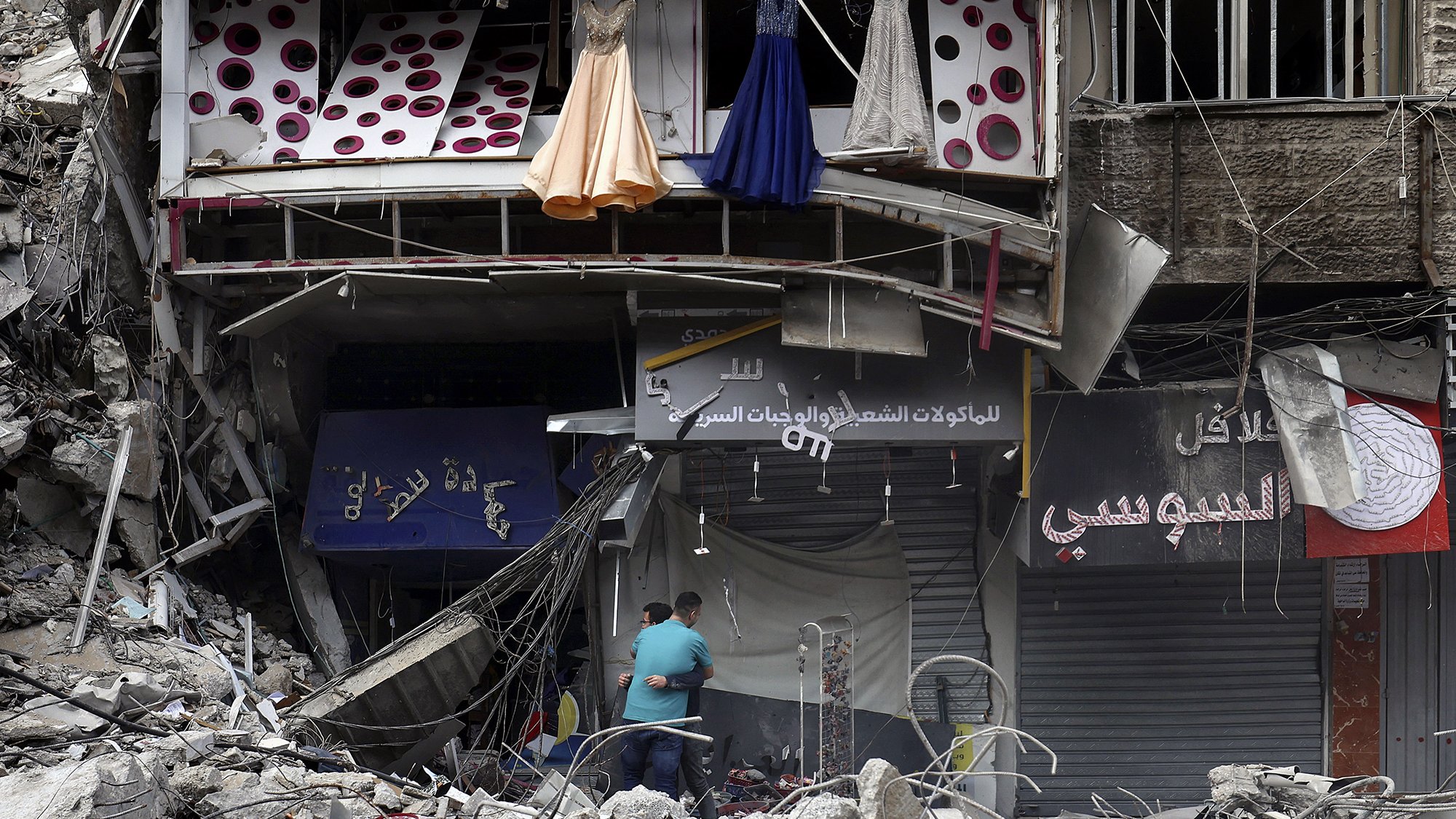 L'image montre des débris d'un bâtiment endommagé, probablement en raison d'un conflit ou d'une catastrophe. À l'avant, on peut voir des robes suspendues à l'extérieur d'une boutique, tandis qu'en bas, deux personnes semblent fouiller ou inspecter les lieux. Les murs sont en ruine, et l'environnement dégage une impression de destruction et de désolation. Des enseignes de magasins sont visibles, mais partiellement masquées par les débris.