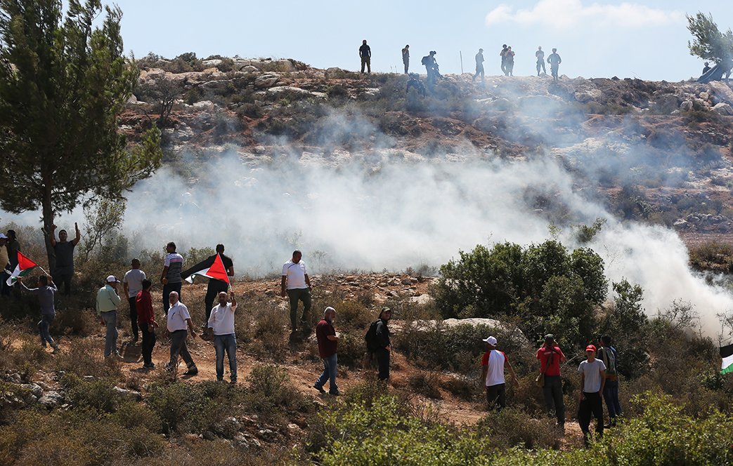 L'image montre une scène de manifestation en plein air. Au premier plan, plusieurs personnes portent des drapeaux palestiniens, tandis que d'autres semblent être en train de marcher ou de s'arrêter. Il y a une atmosphère de tension, avec de la fumée s'élevant dans l'air, probablement due à des gaz lacrymogènes ou à un autre type de dispersion de foule. En arrière-plan, on aperçoit des personnes positionnées sur une colline, qui semblent observer la situation. Le paysage est naturel, avec des arbres et des terrains rocheux.