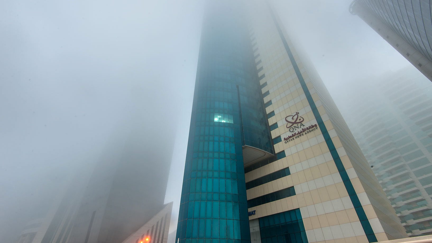 L'image montre un paysage urbain avec des gratte-ciels partiellement cachés par un épais brouillard. La visibilité est réduite, ce qui donne une atmosphère mystérieuse et peu lumineuse. Les bâtiments ont des façades modernes, avec des reflets de verre. On peut distinguer des éléments architecturaux, mais l'intensité du brouillard rend difficile l'observation claire des détails. Les lumières des rues sont visibles, créant un contraste avec l'environnement brumeux.